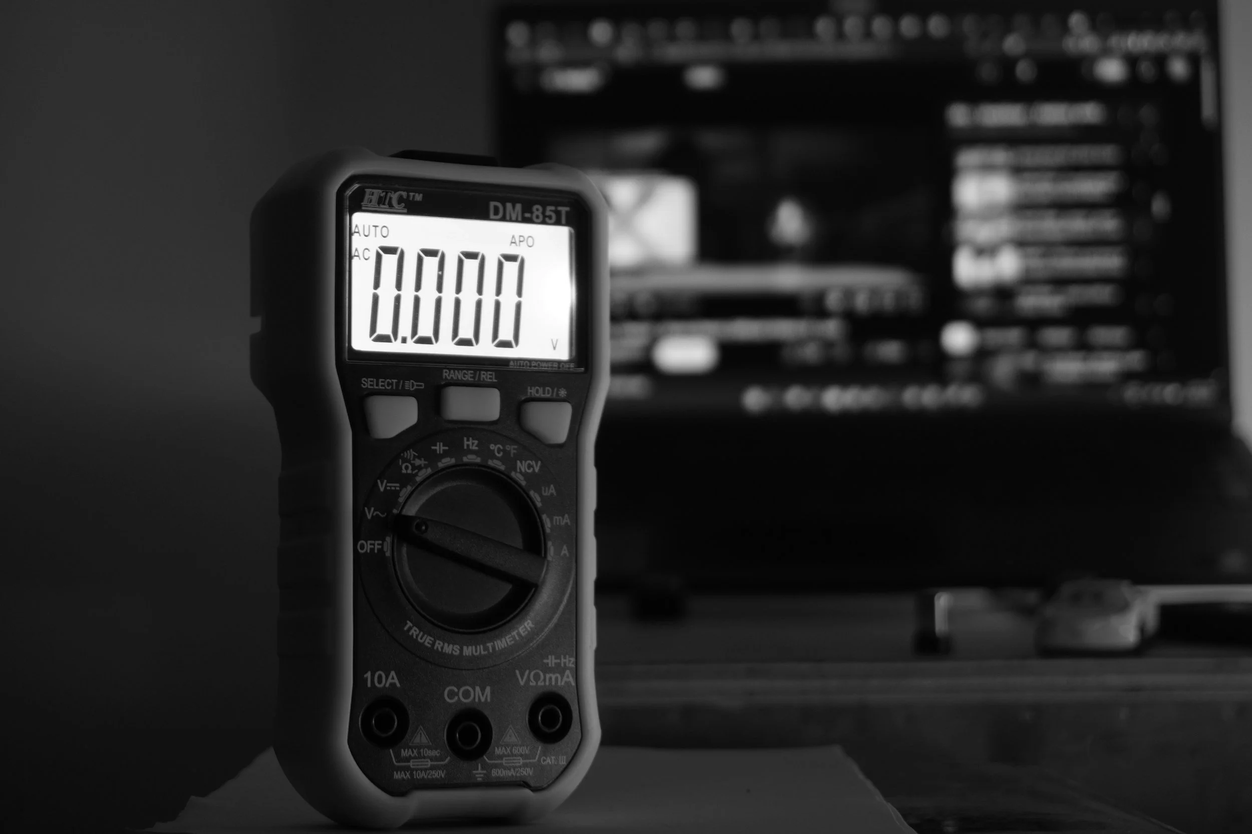 A digital multimeter displaying a reading of 0.000 with a blurred background of electronic equipment.