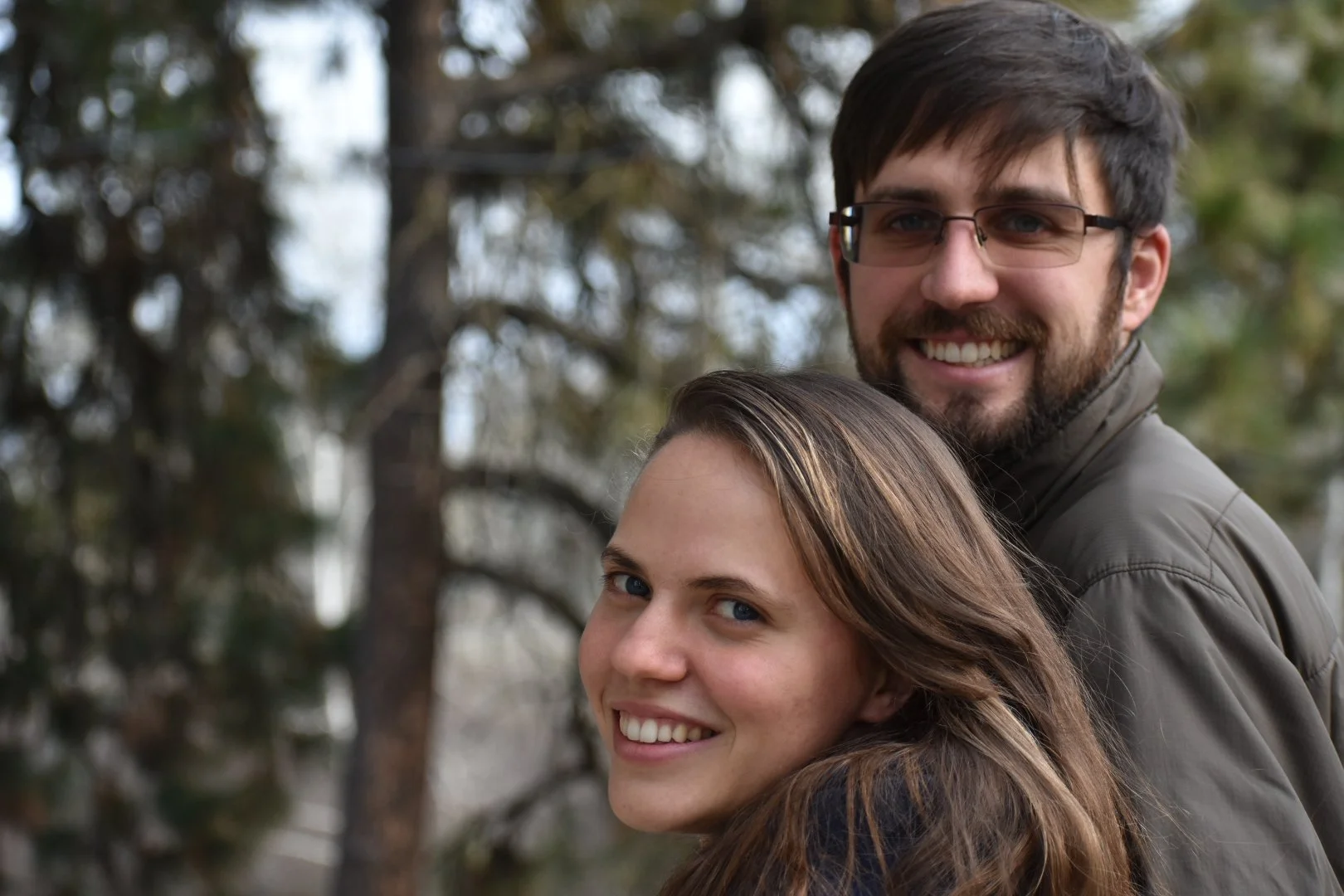 A smiling couple with the woman in front and the man behind, in an outdoor setting with trees in the background.
