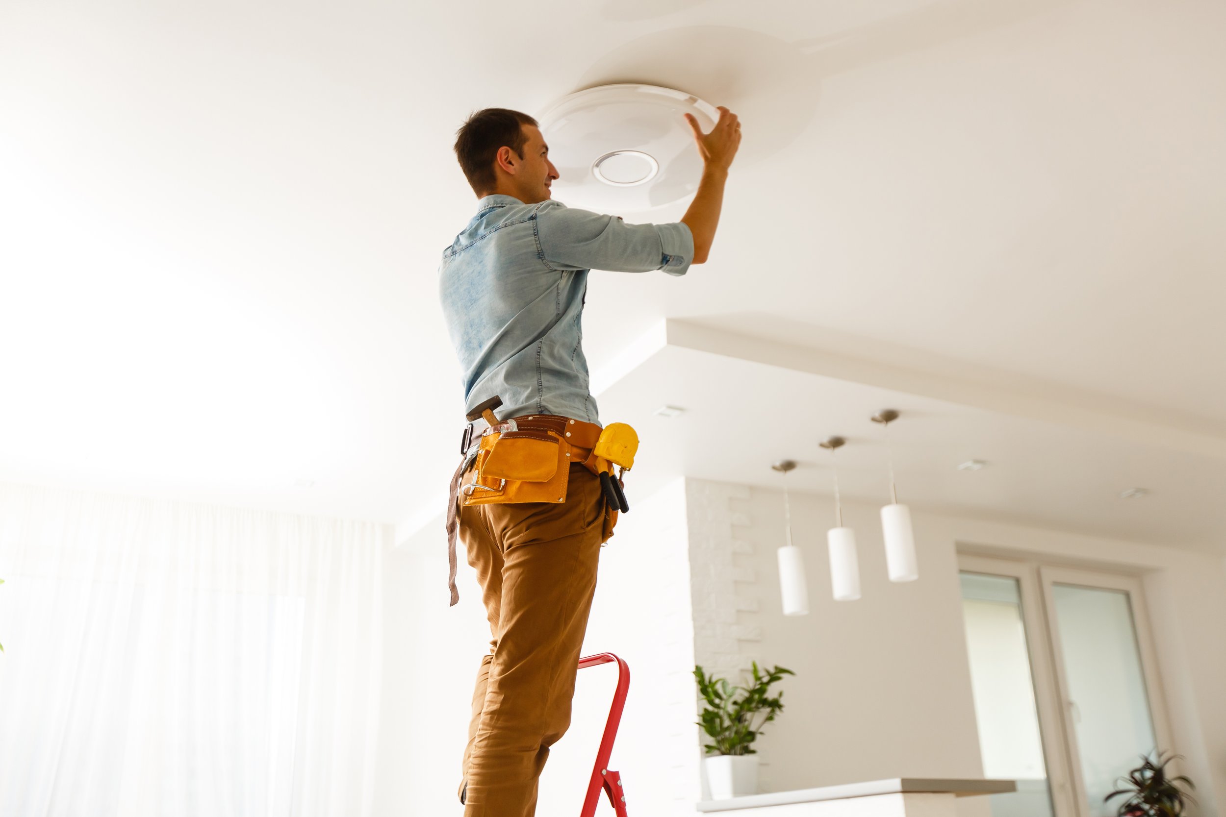 A man in a light denim shirt and brown work pants with a tool belt is installing a ceiling light fixture in a modern, bright living room with a white ceiling and walls, and pendant lights hanging from the ceiling.