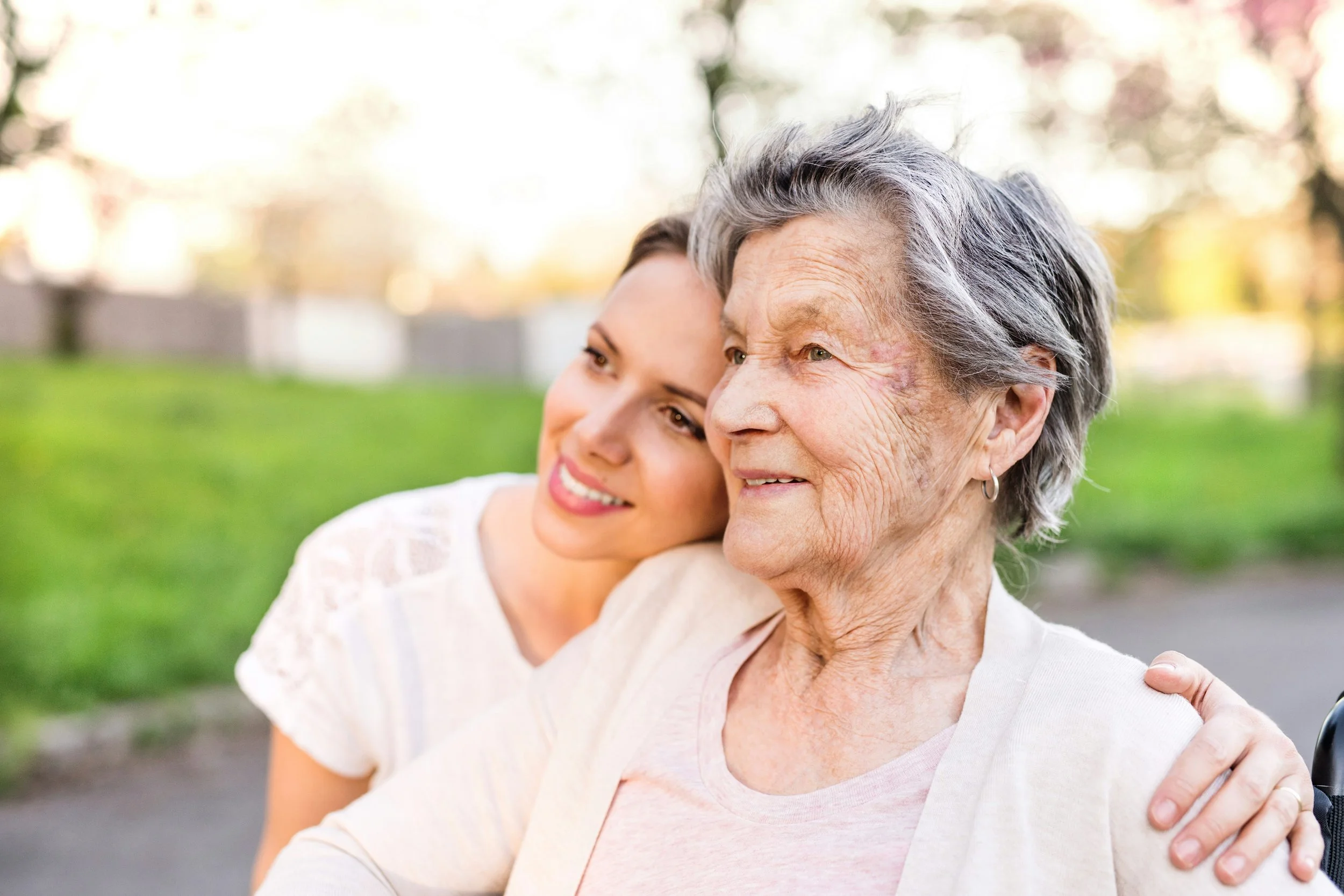 A young woman with brown hair and an older woman with gray hair sharing a hug outdoors amidst trees and greenery.