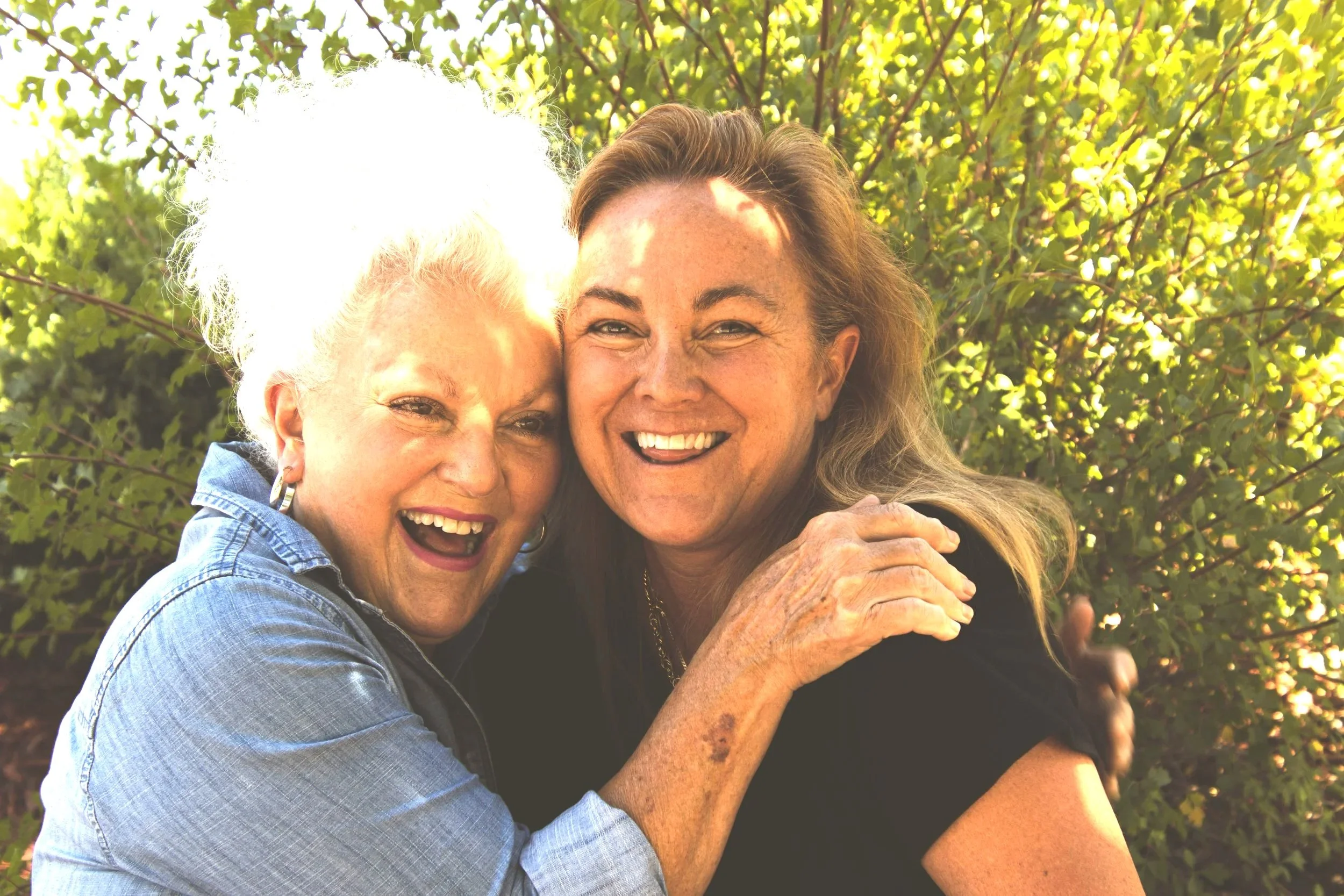 Two smiling women, one with white curly hair and the other with long brown hair, embracing outdoors with green foliage in the background.