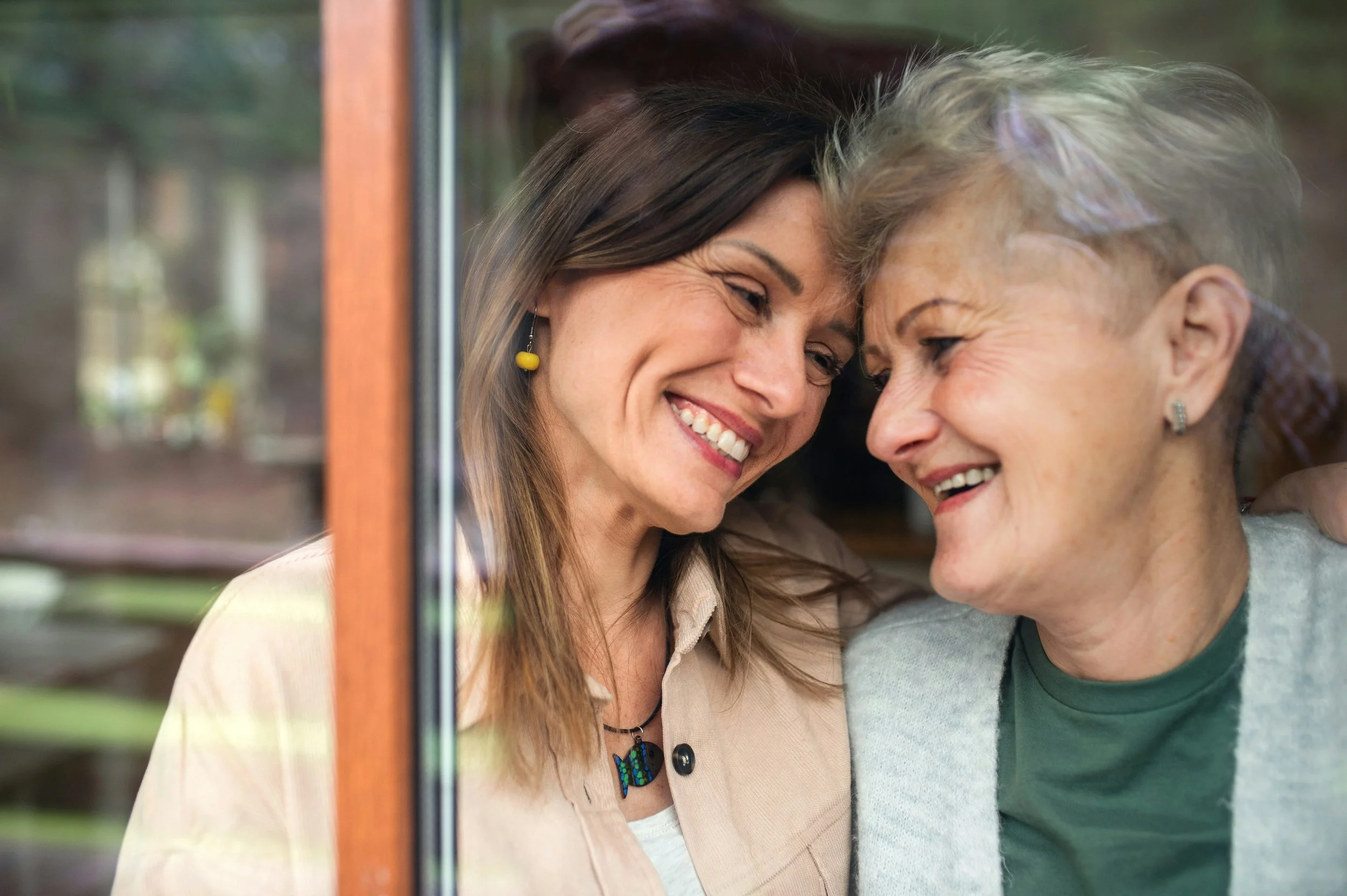 Two women with their foreheads touching, smiling and sharing a tender moment through a glass window.