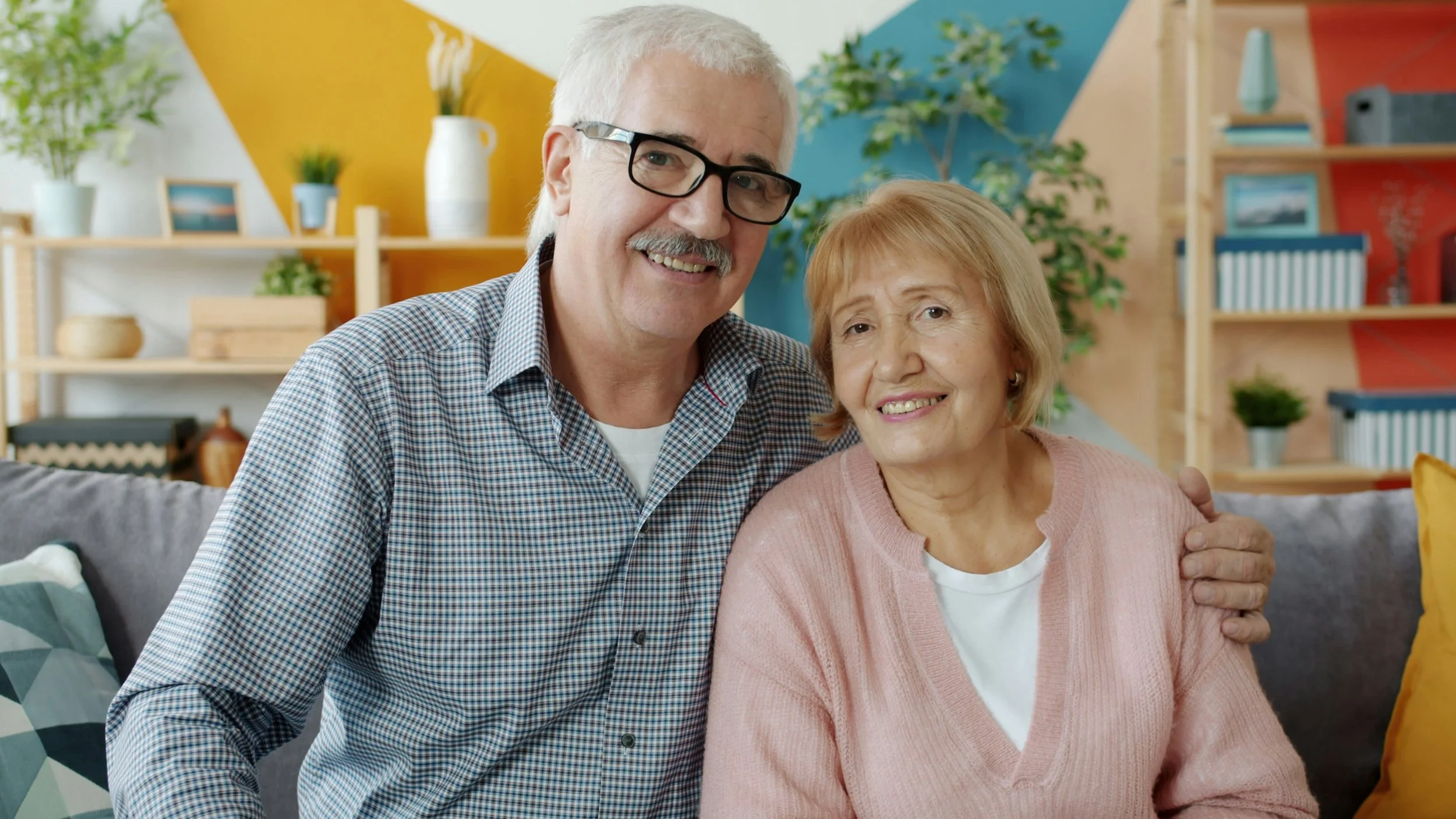 An elderly couple sitting close together on a gray couch in a brightly decorated living room. The man, with white hair and glasses, is smiling and has his arm around the woman. The woman, with short blonde hair, is also smiling. The background includes shelves with plants, books, and decorative items, and a colorful wall with geometric patterns.