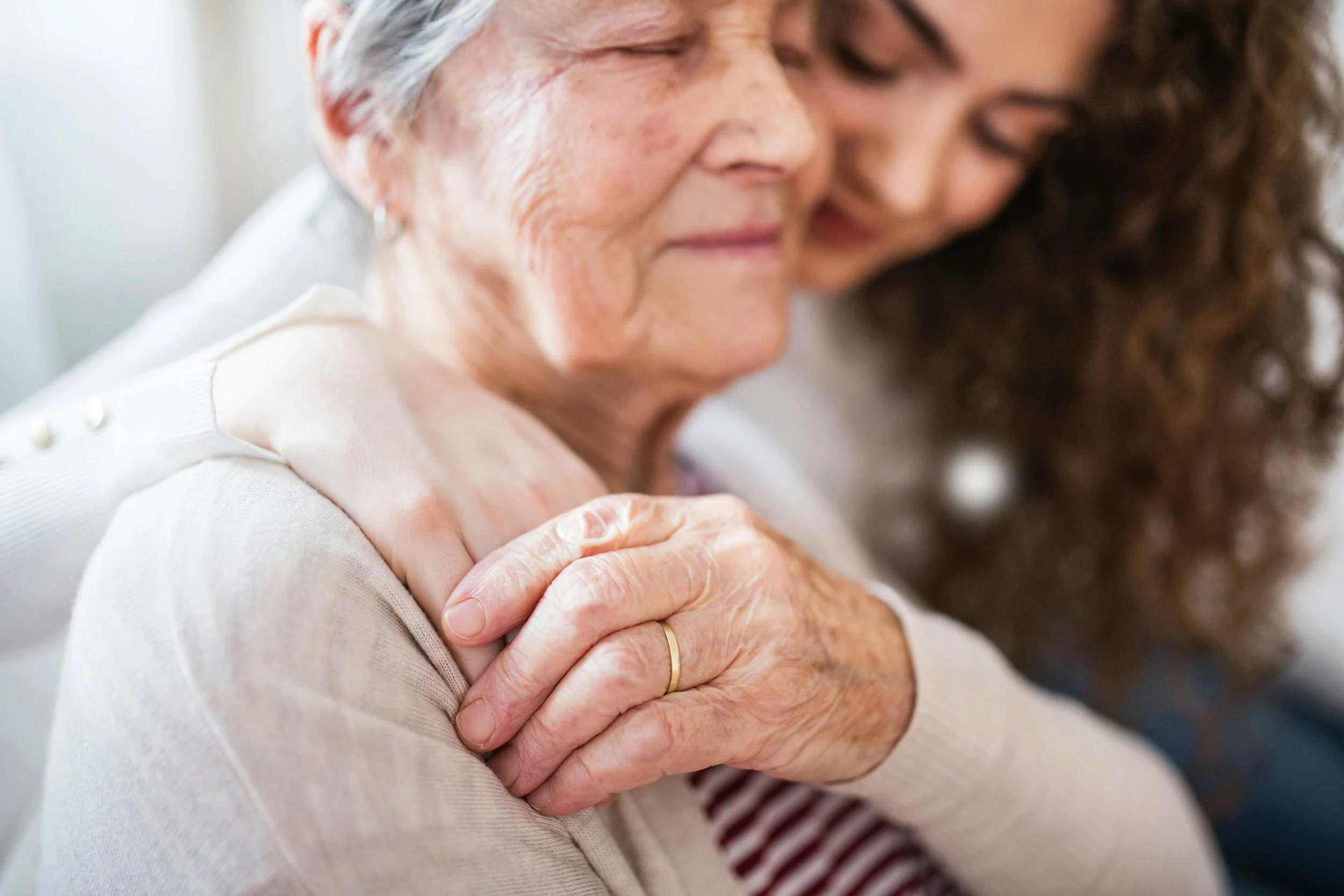 An elderly woman with gray hair is being embraced and comforted by a younger woman with curly hair, both with their eyes closed.