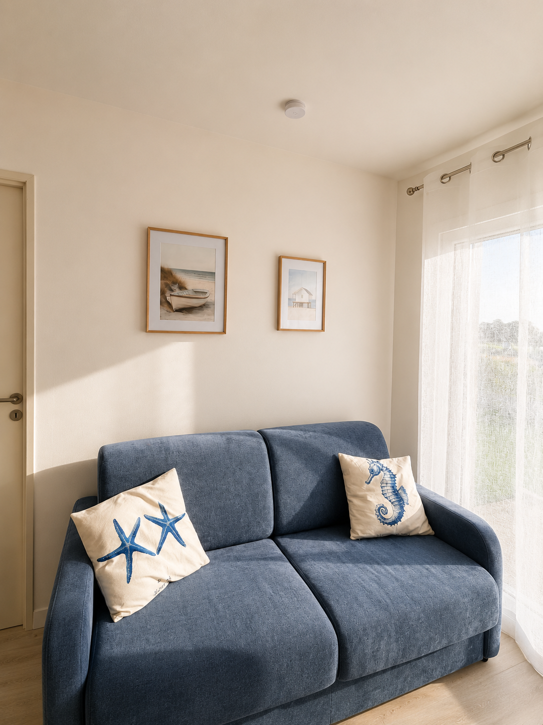 Salon avec un canapé bleu, deux coussins avec motifs de starfish et seahorse, au mur deux tableaux, et une fenêtre à rideaux blancs laissant entrer la lumière.