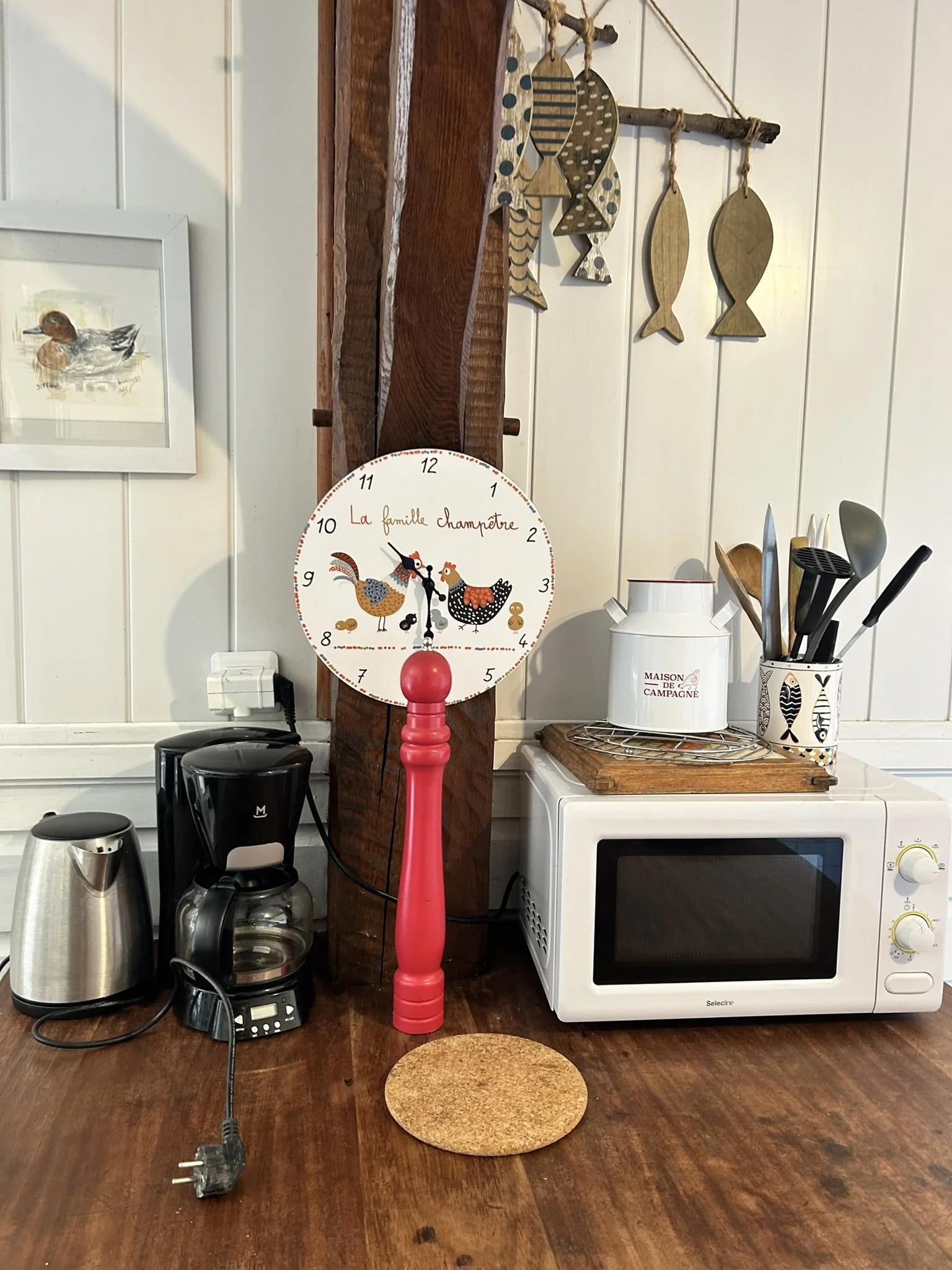 Une cuisine avec un micro-ondes blanc, une cafetière électrique, un thermos en acier inoxydable, un porte-ustensiles avec des cuillères et une spatule, uniquement la partie inférieure d'une horloge avec des dessins de poules, une planche à découper ronde en liège, et un mécanisme électrique branché sur une prise électrique.