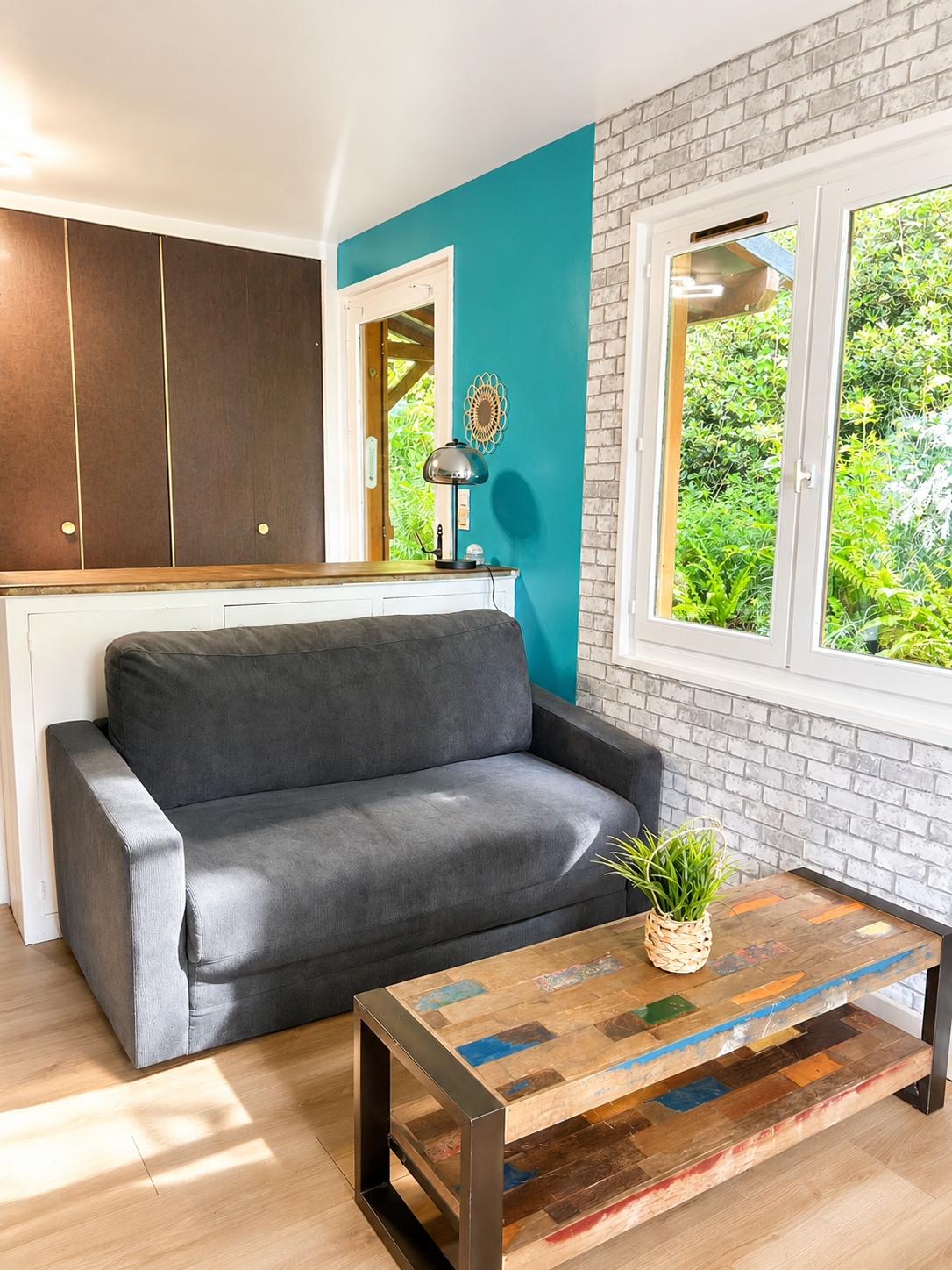Salon avec canapé gris, table en bois, mur en briques blanches, fenêtre donnant sur un jardin verdoyant, lampe de bureau, décoration murale.