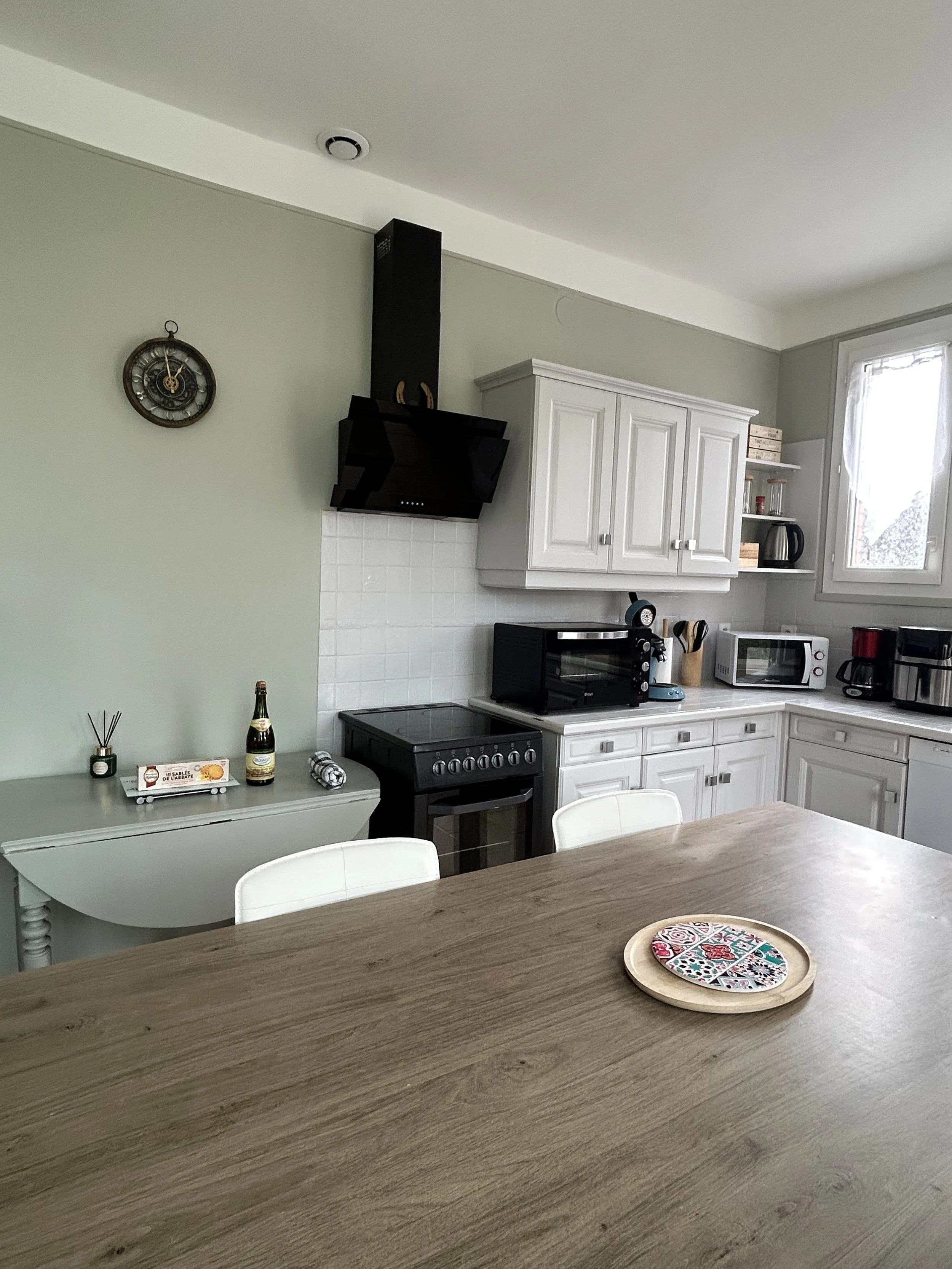 Cuisine moderne avec meubles blancs, un four, un micro-ondes, une cafetière, un plan de travail en bois clair, et un table avec des chaises blanches. Un plateau avec un motif coloré est sur la table.