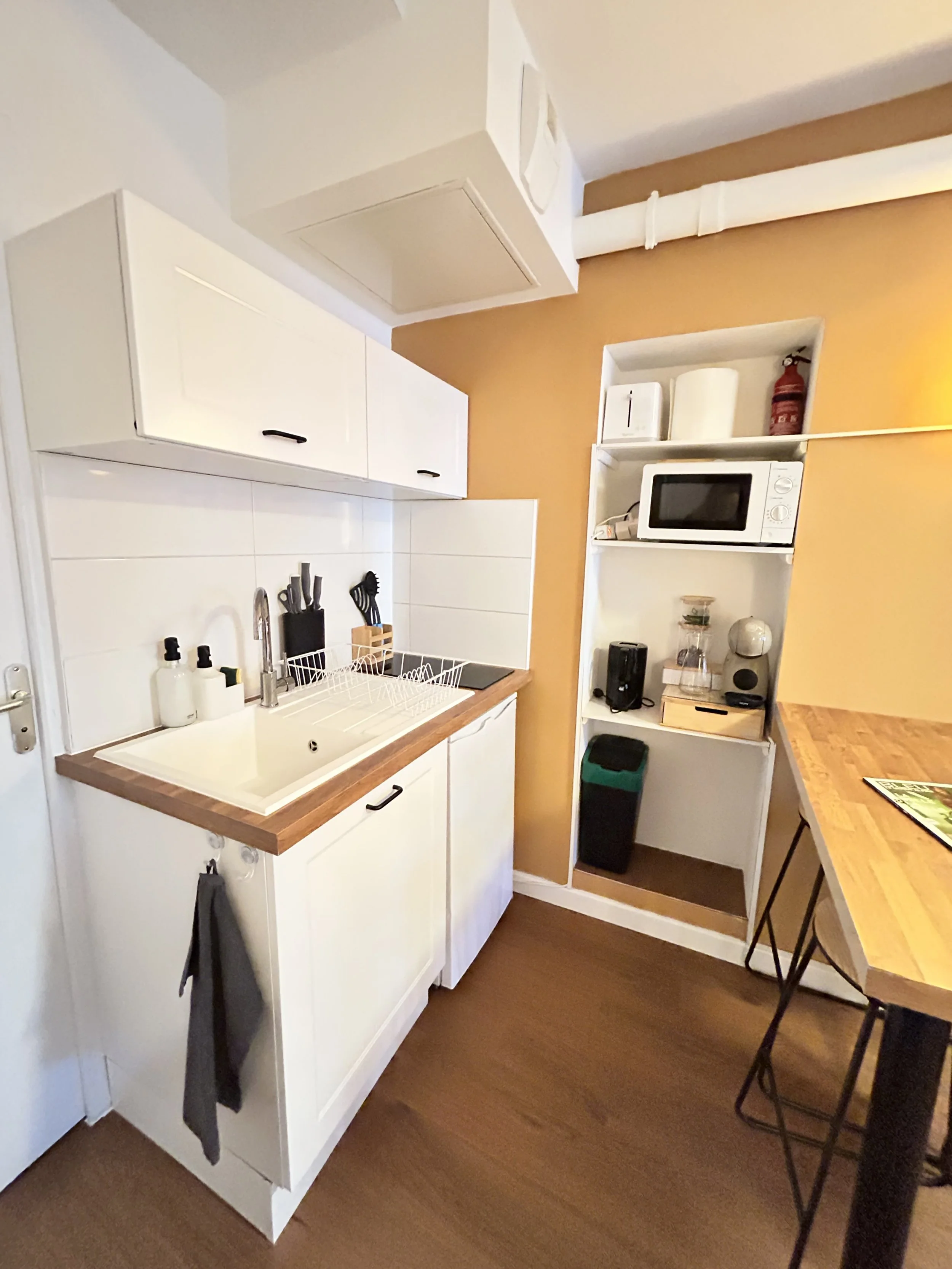 Une cuisine moderne avec un évier, des étagères avec des appareils électroménagers et des ustensiles, une table en bois avec des chaises hautes en métal, et un mur peint en orange.