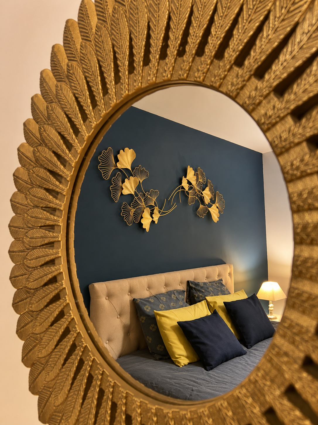 Reflet dans un miroir orné de motifs en forme de feuilles dorées, montrant un lit avec des coussins colorés et une décoration murale en métal représentant des feuilles, dans une chambre avec une lumière douce.