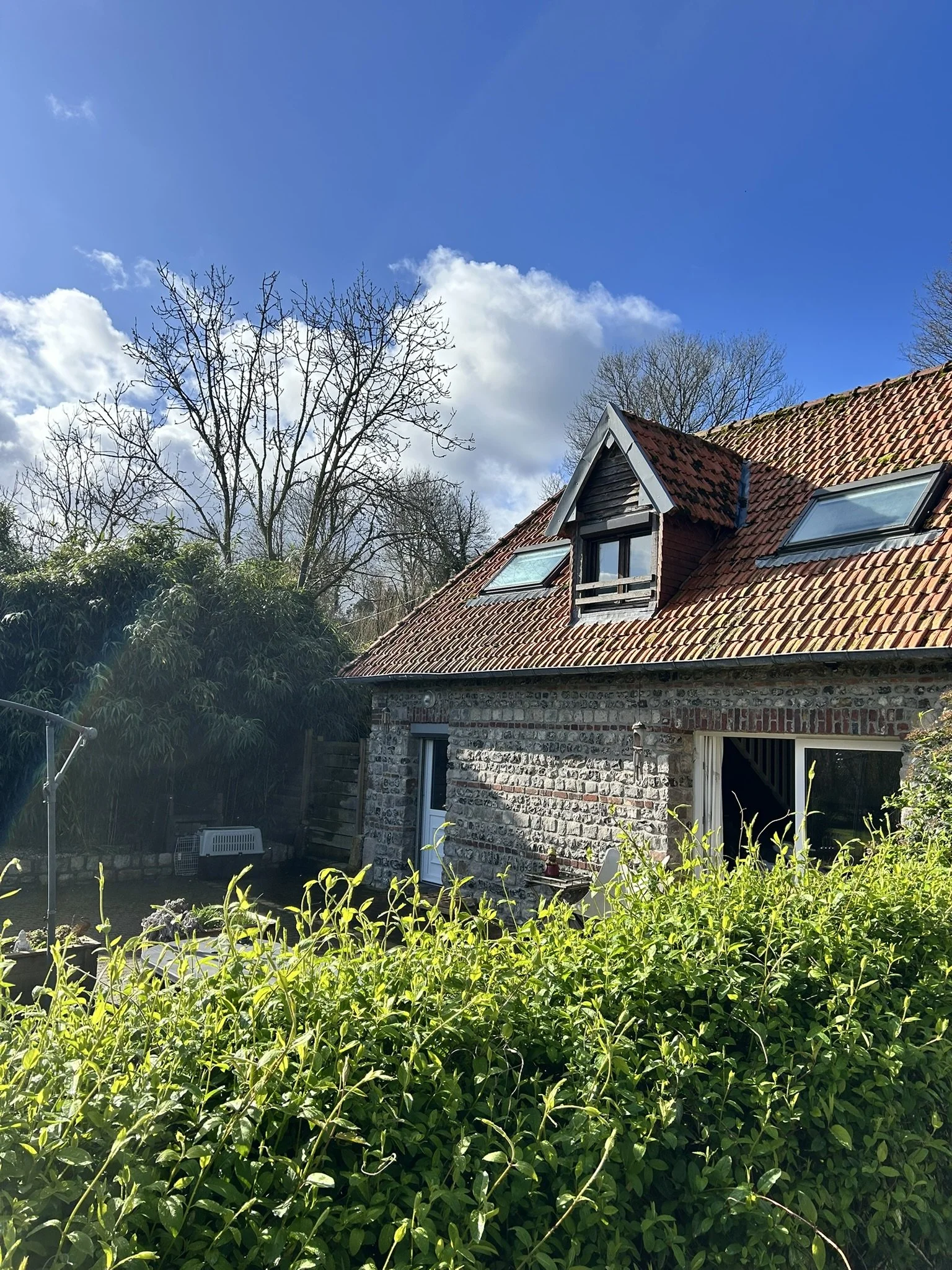 Maison en pierre avec toiture en tuiles rouges, fenêtres de toit, jardin avec végétation dense, arbres sans feuilles, ciel bleu avec quelques nuages.