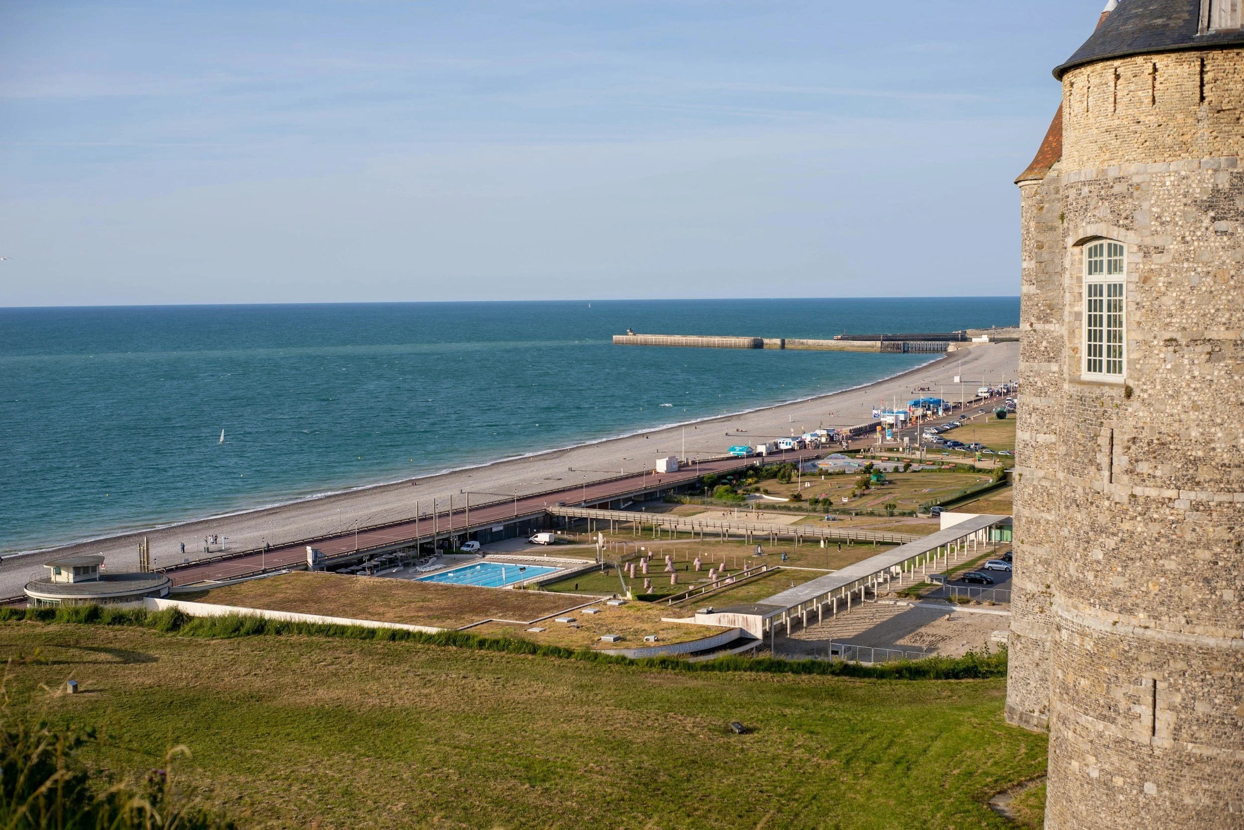 Vue d'une côte avec un bâtiment en pierre à droite, une plage, un front de mer et la mer au fond.