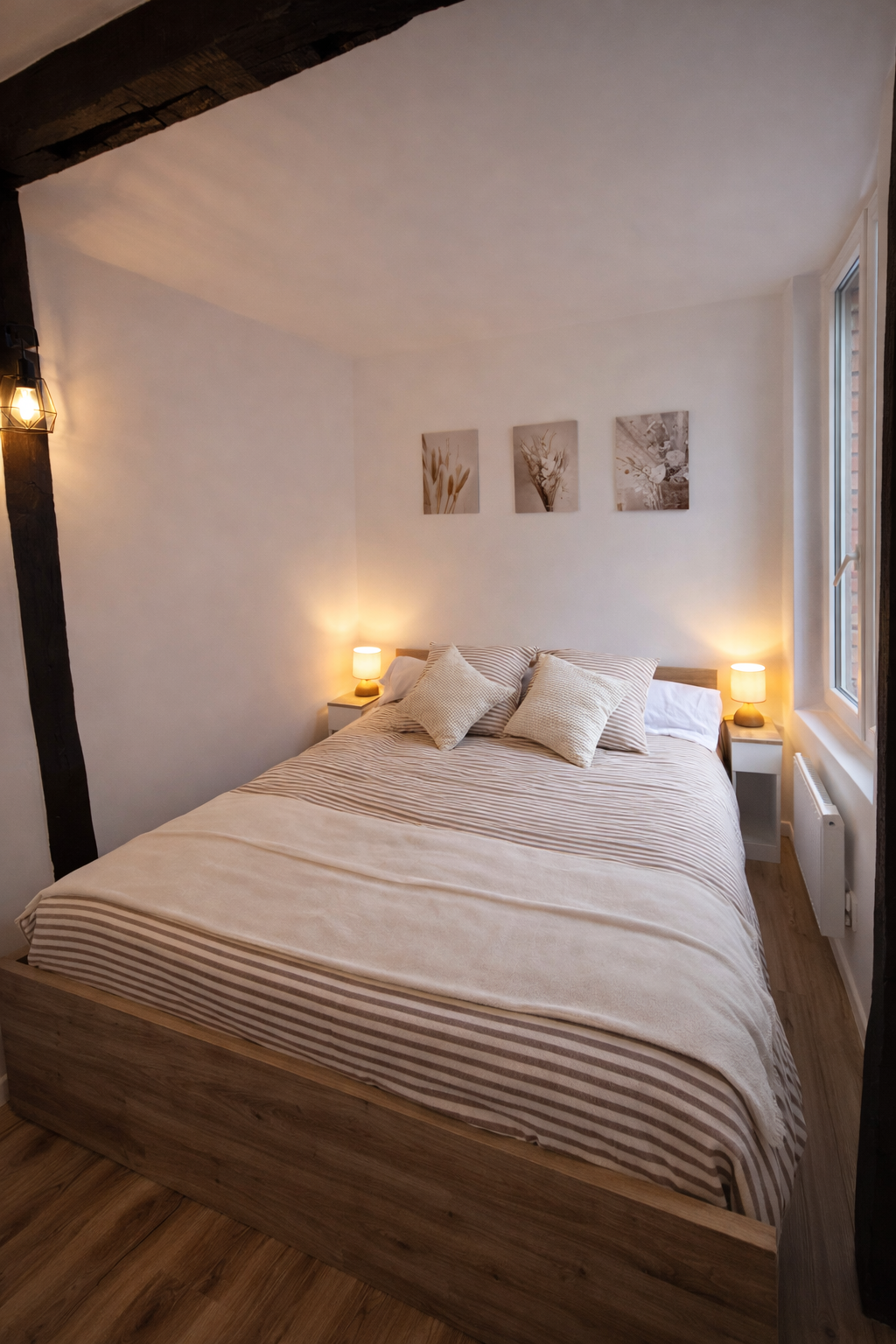 Chambre avec lit king-size avec draps à rayures beige et blanc, trois oreillers, deux tables de nuit avec lampes, décor minimaliste, fenêtre lumineuse, murs blancs, plafond en bois.
