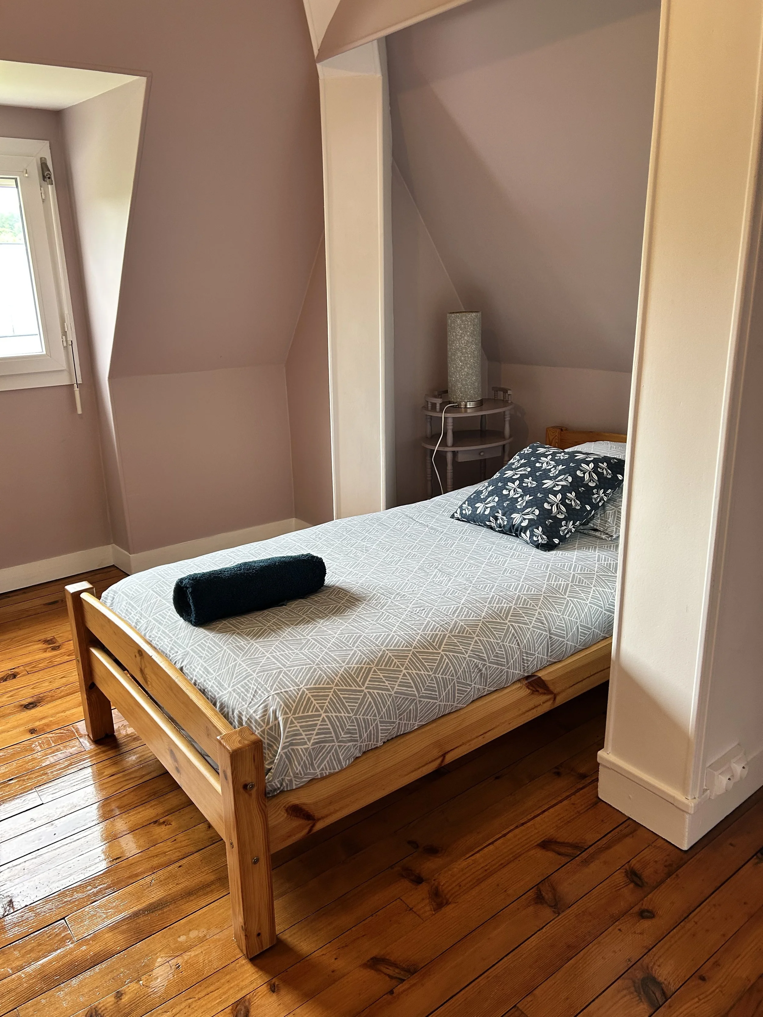 Un lit en bois dans une chambre avec parquet en bois. La tête de lit est en bois, avec un oreiller à motif floral noir et blanc. À côté du lit, une petite table blanche avec une lampe cylindrique et un drap noir roulé sur le lit.