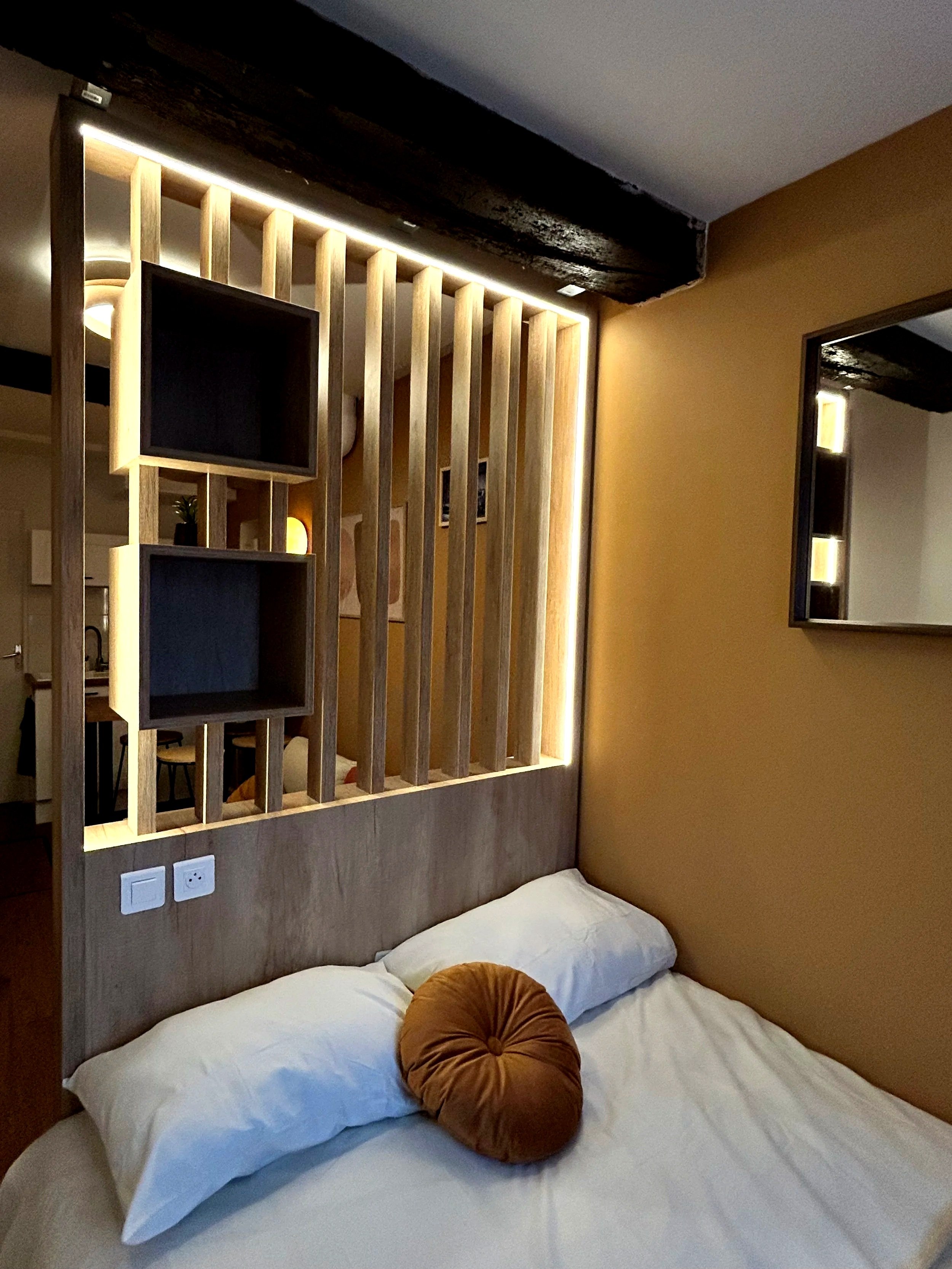 Lit avec des oreillers blancs et un coussin rond brun, tête de lit en bois avec un panneau séparateur en bois décoratif, deux téléviseurs encastrés, miroirs, lampes, et murs en couleur beige