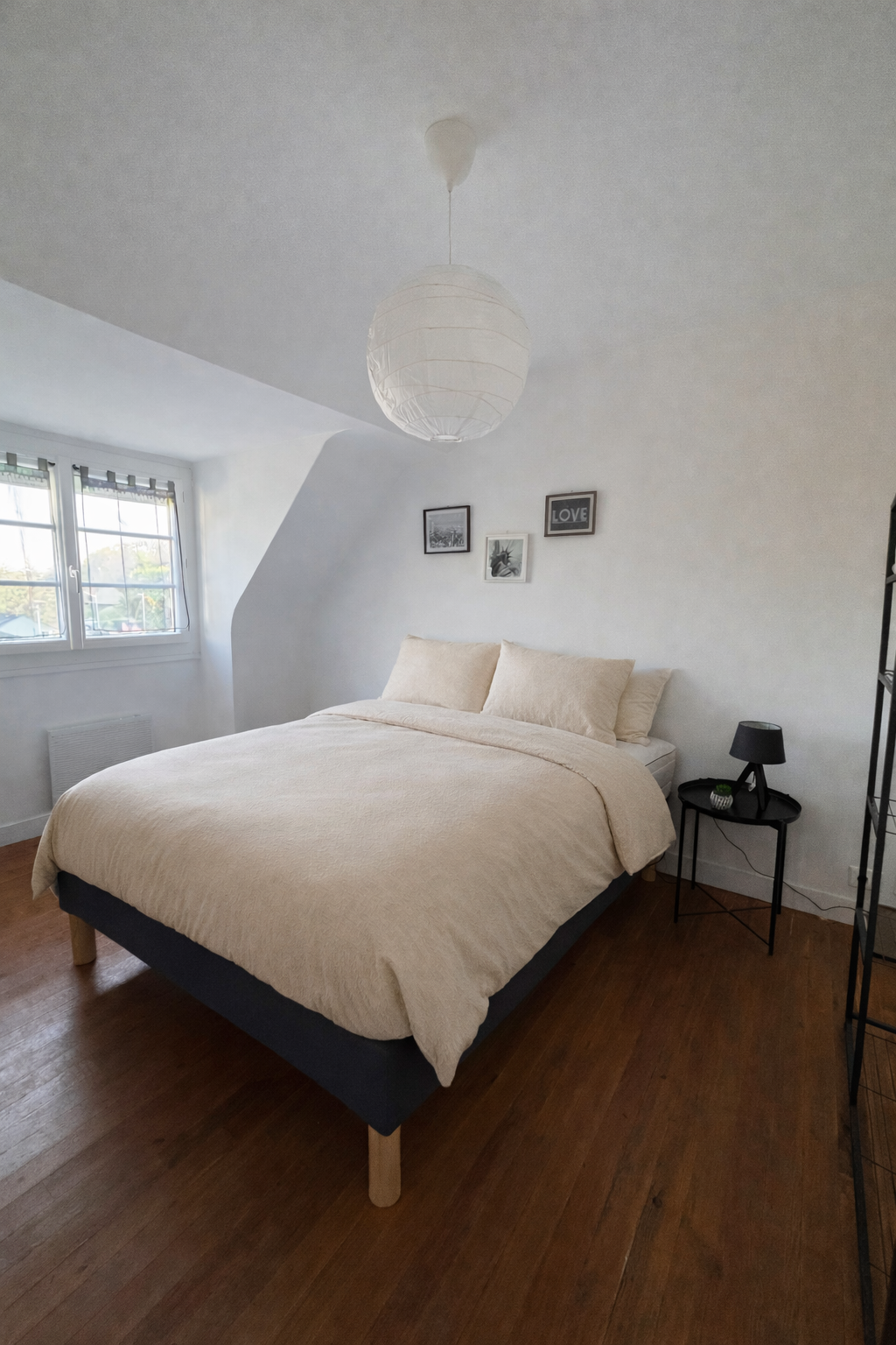 Chambre avec lit double, draps beiges, tableau noir et blanc, cadre avec le mot 'LOVE', lampe de chevet noire, fenêtre avec vue sur la nature, sol en bois.