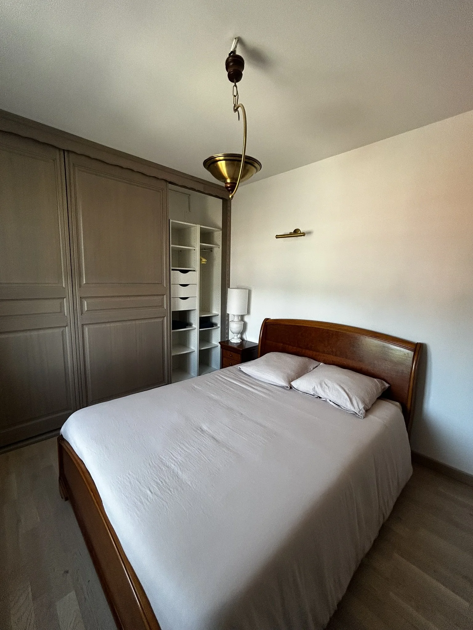 Chambre avec un grand lit en bois, deux oreillers, une armoire grise, une lampe de chevet blanche, un lampadaire et une suspension au plafond.