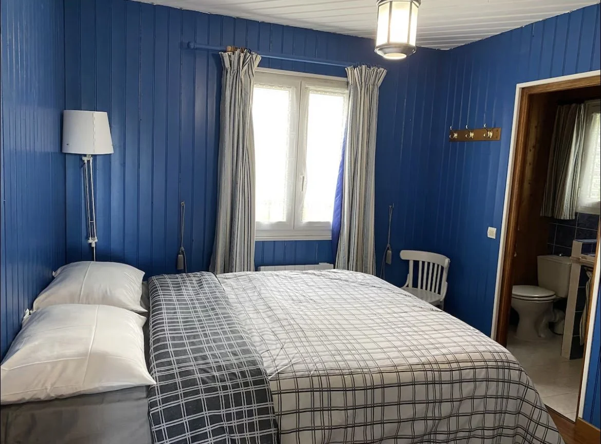 Chambre avec murs en bois colorés en bleu, lit double avec draps à carreaux, fenêtre avec rideaux, lampe suspendue au plafond, petite chaise en bois, porte menant à une salle de bain.