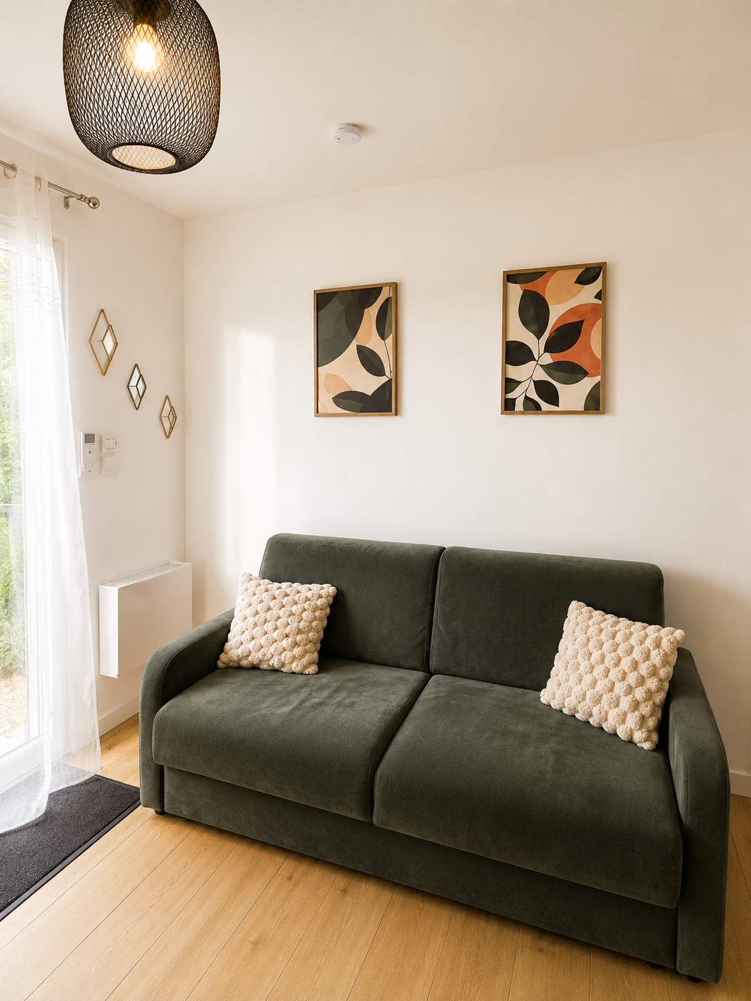 Un salon avec un canapé vert foncé, deux coussins en peluche beige, une peinture murale avec des motifs de feuilles colorées, une lampe suspendue en noir, un sol en bois clair et une porte-fenêtre avec rideaux transparents laissant entrer la lumière.