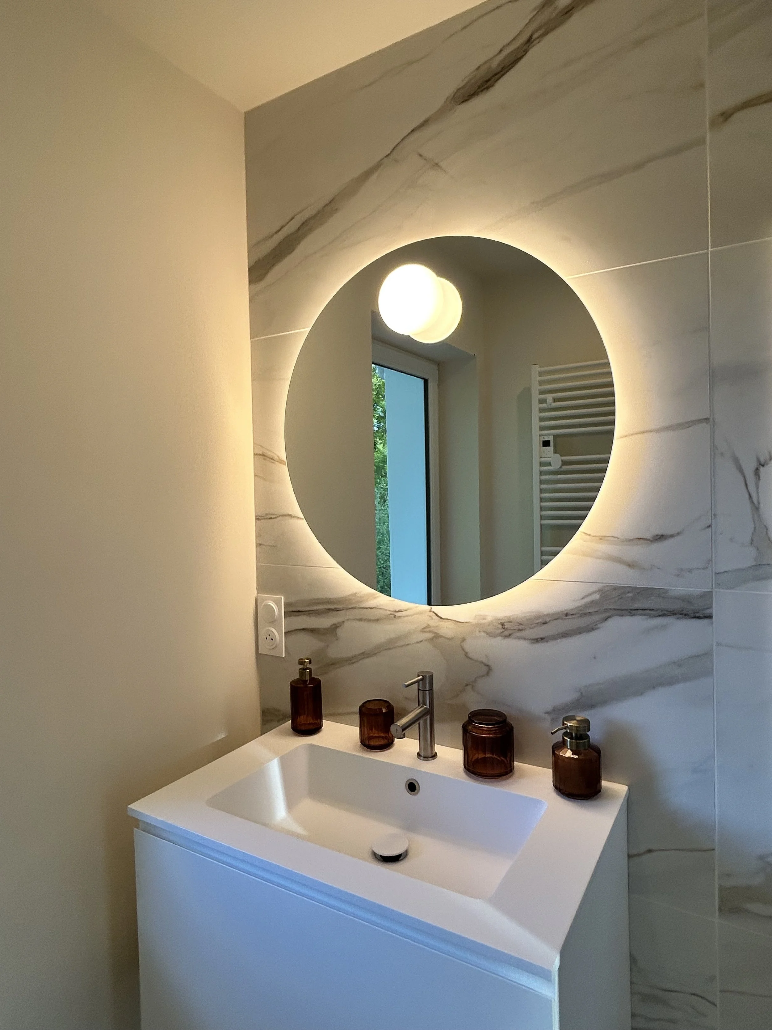 Un miroir rond avec un éclairage LED intégré, un lavabo blanc moderne, des flacons en verre amber sur le comptoir, une fenêtre laissant entrer de la lumière naturelle et un radiateur électrique blanc derrière le miroir dans une salle de bain élégante.