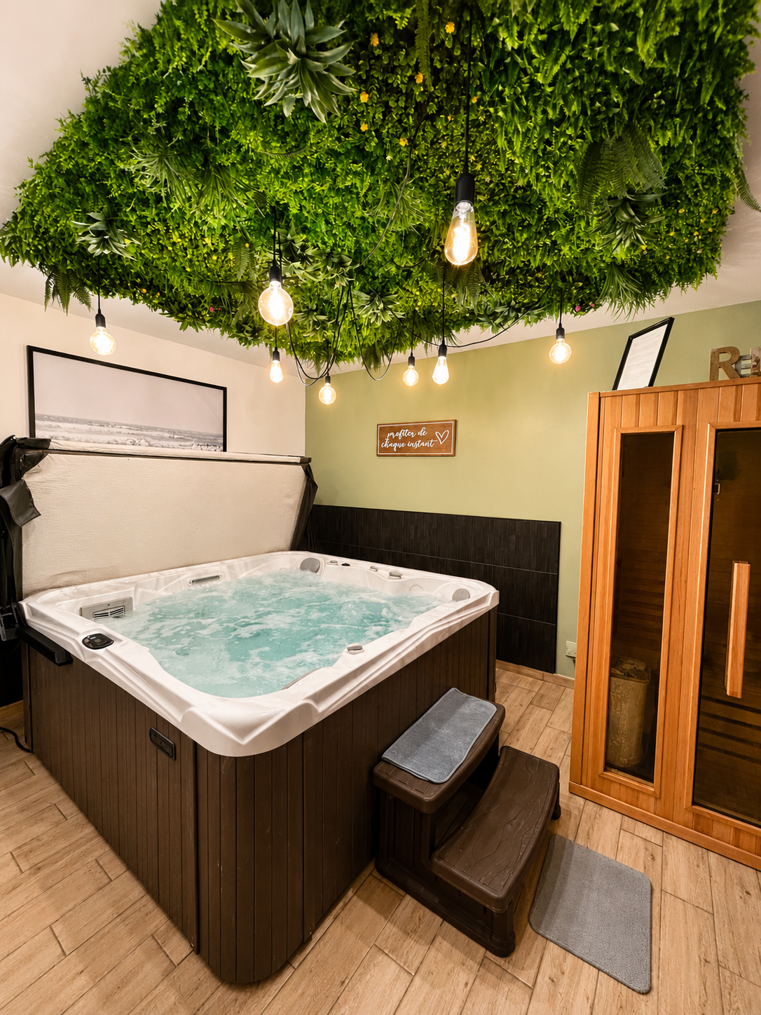 Spa intérieur avec jacuzzi à jets d'eau, sous un ceiling couvert de plantes vertes, éclairé par des ampoules suspendues, dans une pièce avec un sauna en bois et des murs décorés de citations.