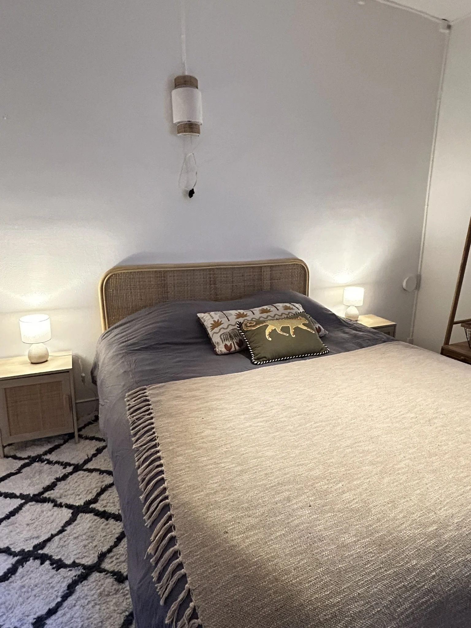 Chambre avec un lit à baldaquin, deux lampes de chevet, un tapis avec motif en zigzag noir et blanc, une tête de lit en rotin, et un coussin décoratif avec un animal en or.