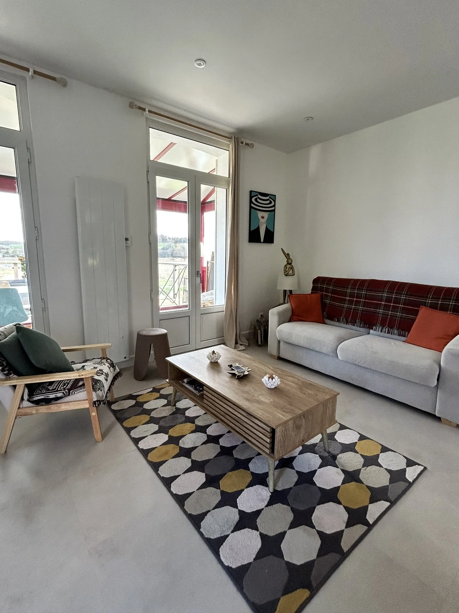 Salon avec un canapé blanc, un tapis géométrique multicolore, une table en bois, une chaise avec un coussin vert, une lampe avec une décoration en forme de lapin, un tableau coloré, et un balcon en arrière-plan.