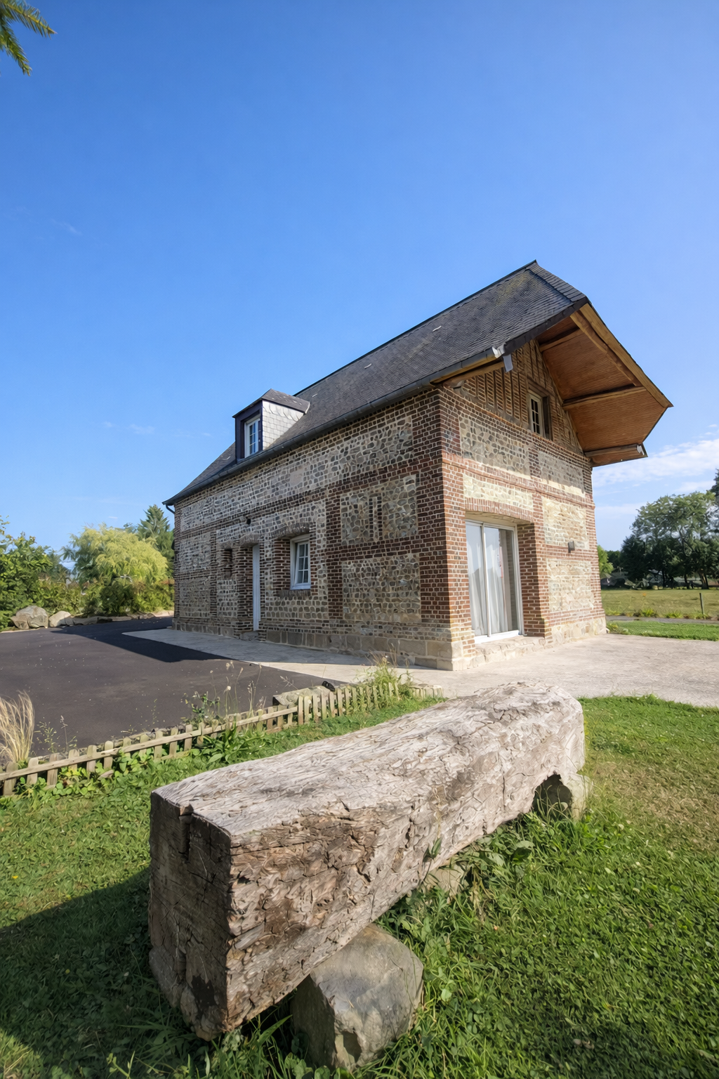Maison en pierre avec une toiture en pente, fenêtres et porte en verre, dans un environnement rural avec un banc en bois devant et un ciel bleu