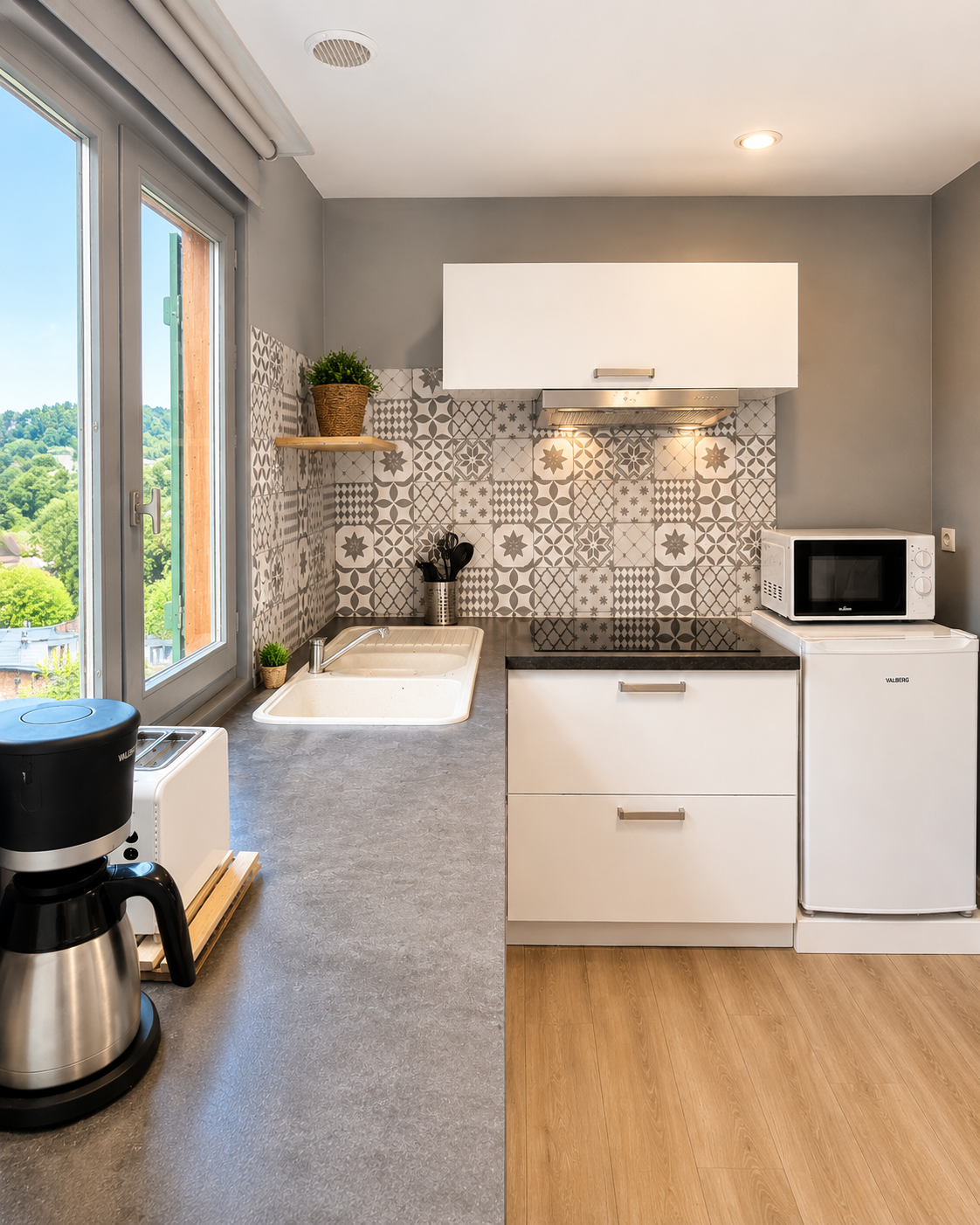 Cuisine moderne avec fenêtre donnant sur un paysage verdoyant, comptoir gris, évier, plaque de cuisson noire, micro-ondes blanc sur réfrigérateur, décoration végétale, carrelage mural à motifs en gris et blanc, ustensiles de cuisine.