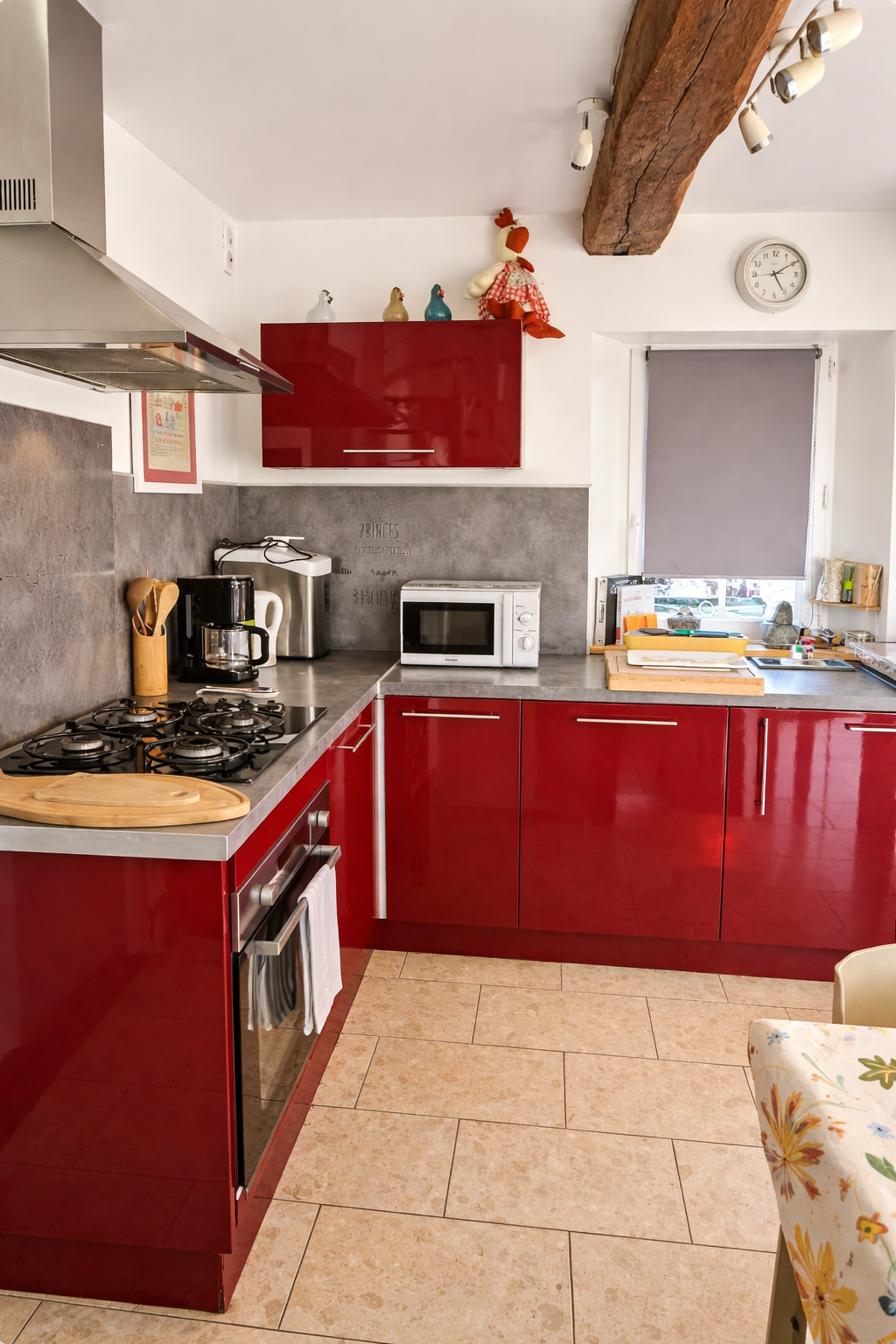 Cuisine moderne avec des armoires rouges brillantes, un four, une cuisinière à gaz, un micro-ondes et des appareils électroménagers, un plan de travail en pierre, une fenêtre avec un volet gris, decorations sur l'étagère supérieure et le plafond en bois.