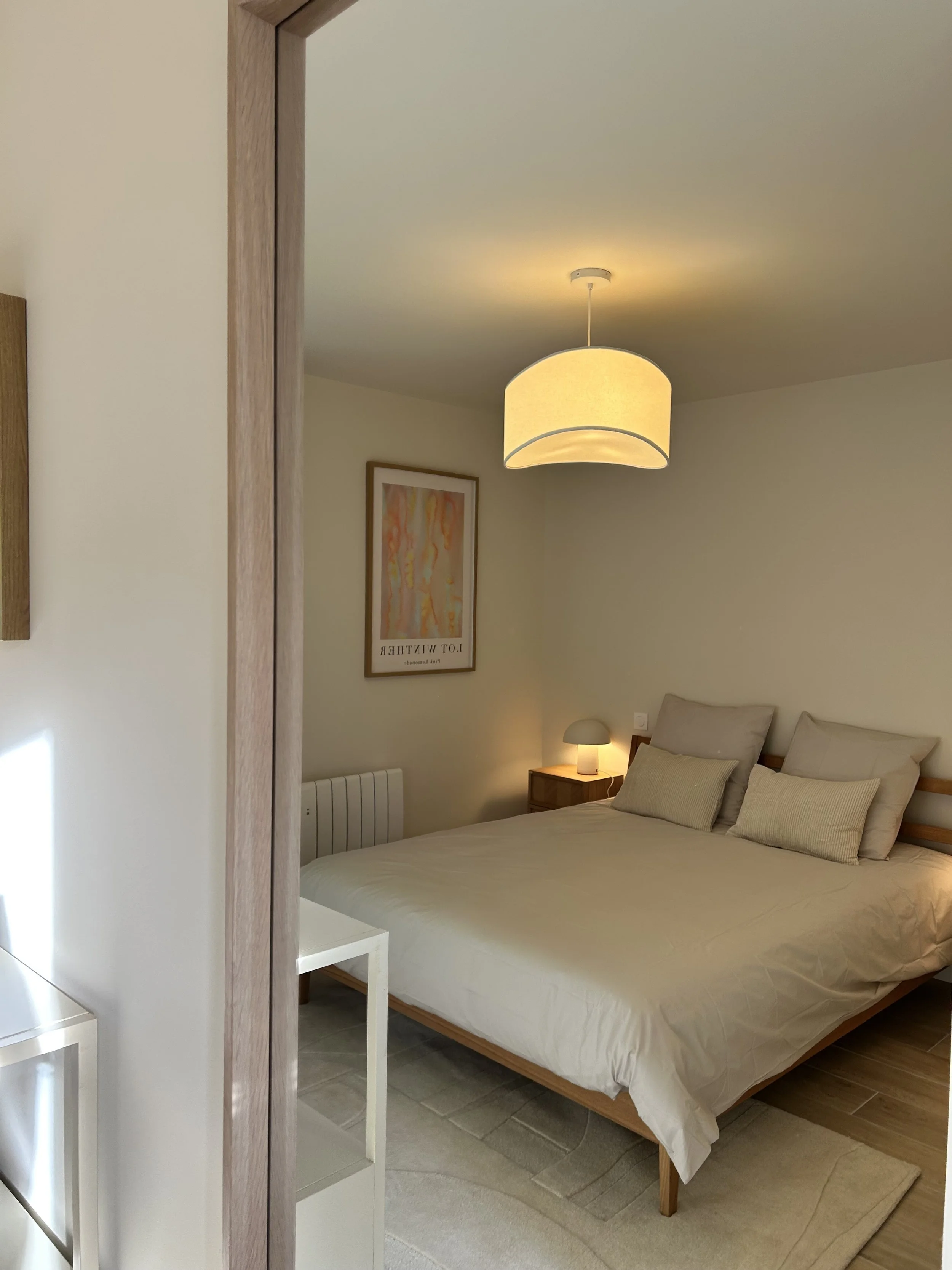 Photo d'une chambre avec un lit double, des coussins beiges, une petite table de chevet avec une lampe, une œuvre d'art encadrée sur le mur, un plafond avec un luminaire suspendu