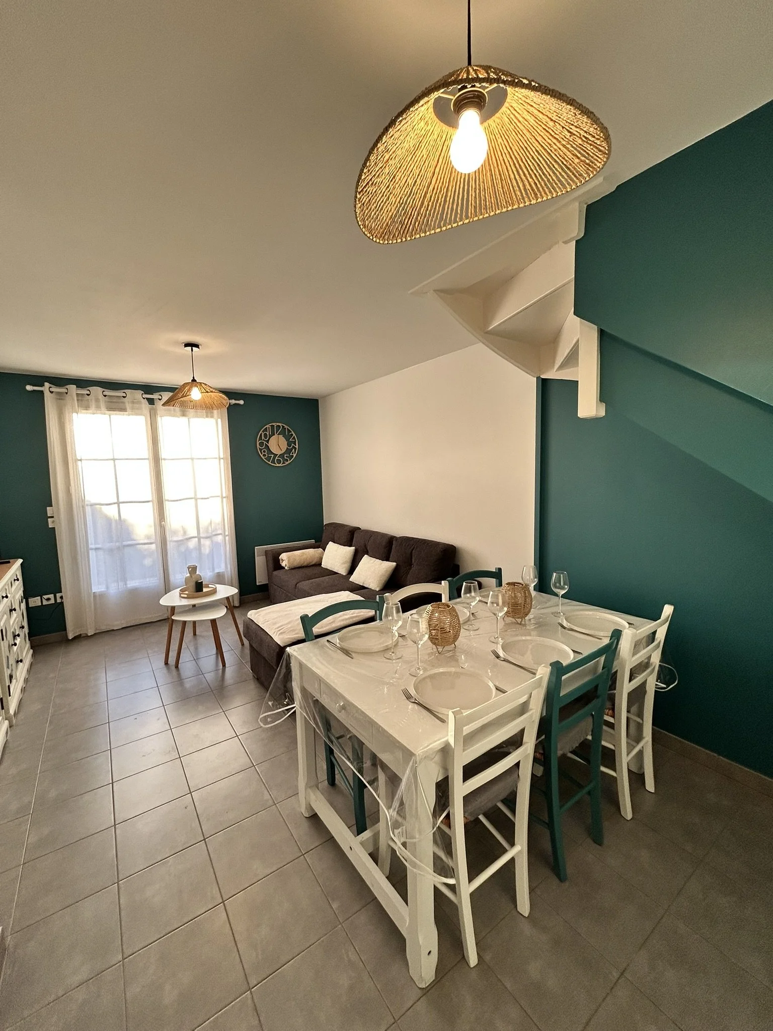 Salon intérieur avec un canapé gris, une table basse, des rideaux blancs devant une fenêtre, un mur vert avec une horloge, un plafond avec deux lampes suspendues à abat-jour en rotin, et une table de salle à manger avec des chaises, des verres et des assiettes.
