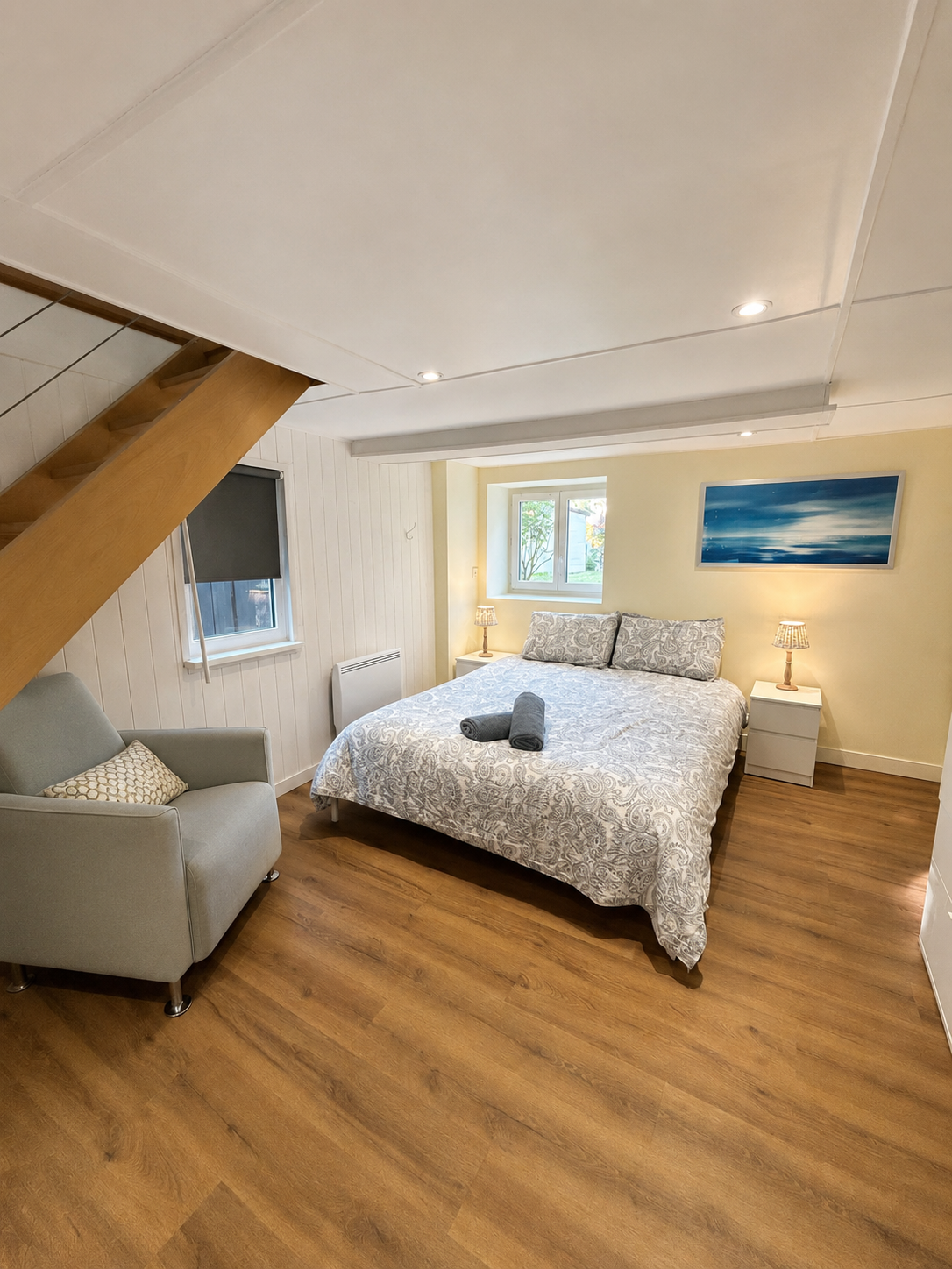 Chambre avec lit double, deux lampes de chevet, tableau abstrait et fauteuil gris.