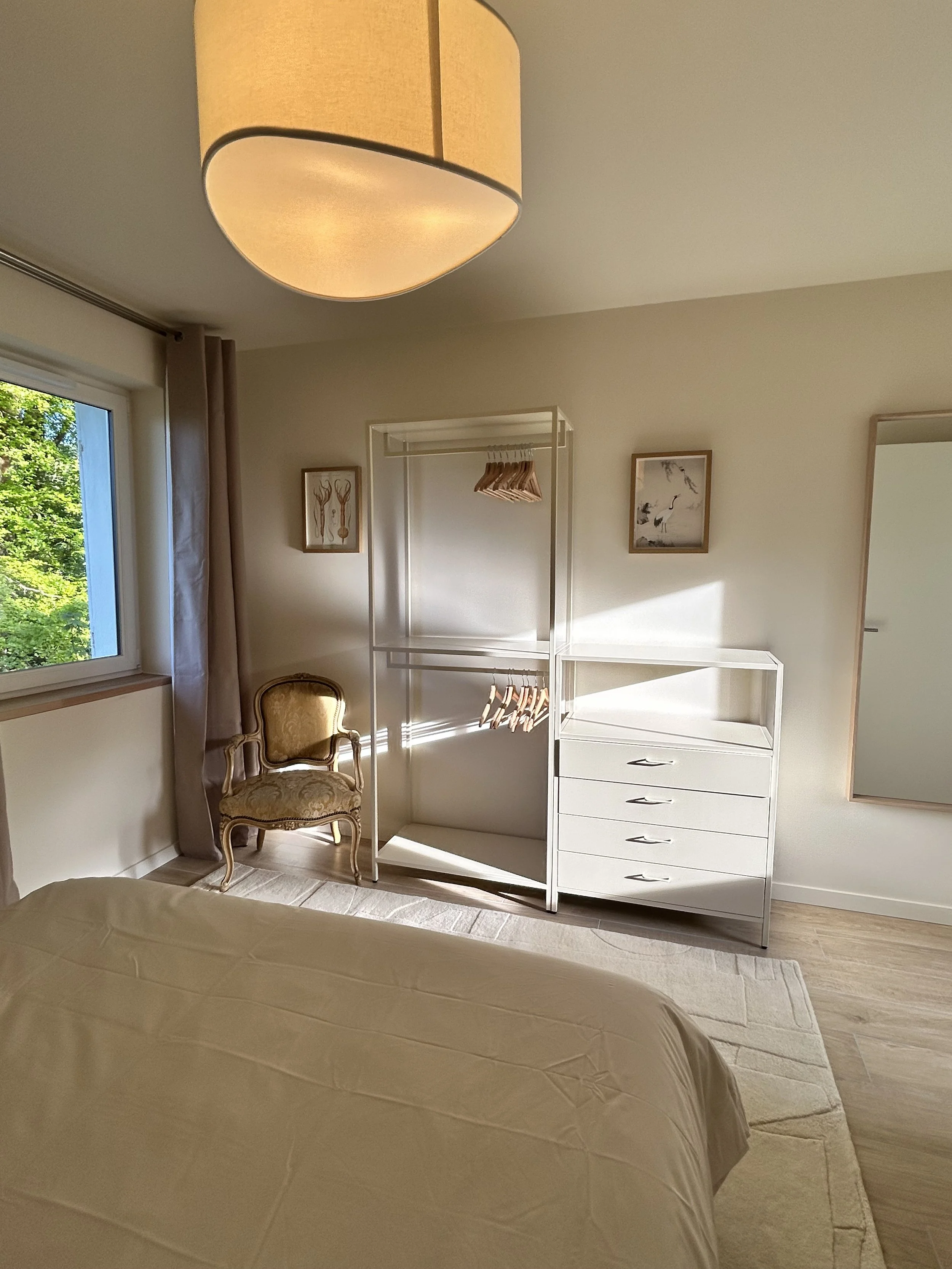 Vue partielle d'une chambre avec un lit, une fenêtre avec rideaux, un fauteuil ancien, un meuble blanc avec tiroirs, des œuvres d'art encadrées et un porte-manteaux vide.