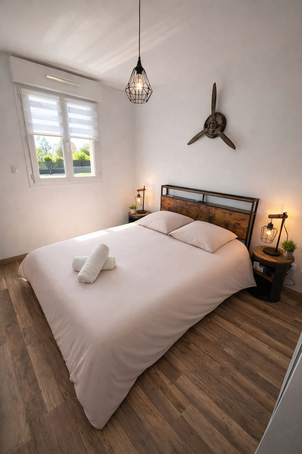 Chambre de style moderne avec un lit double, draps blancs, deux oreillers, et serviettes roulées. Mur blanc avec un mur en bois derrière le lit, deux tables de nuit avec lampes d'appoint, une fenêtre avec volets roulants, un plafond avec une lampe suspendue, décoration murale de rotor de ventilateur en bois, sol en bois clair.