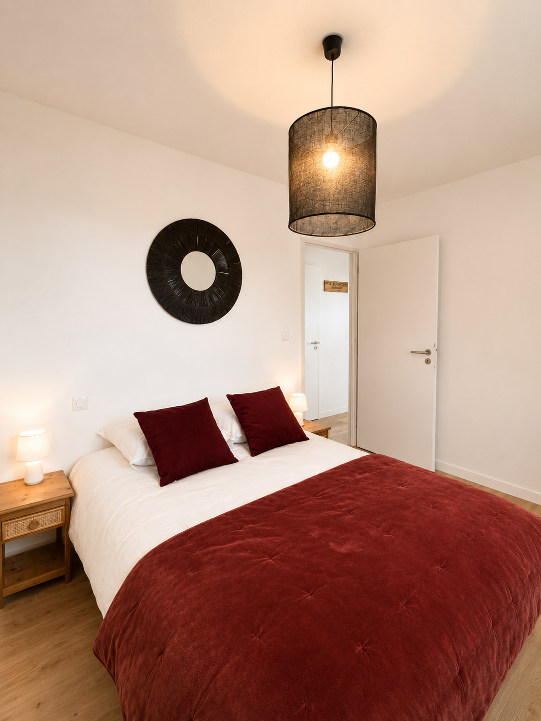 Chambre avec lit double, oreillers rouges, drap de lit rouge, deux lampes de chevet, un mur blanc, un décor noir au mur, et une suspension noire au plafond.
