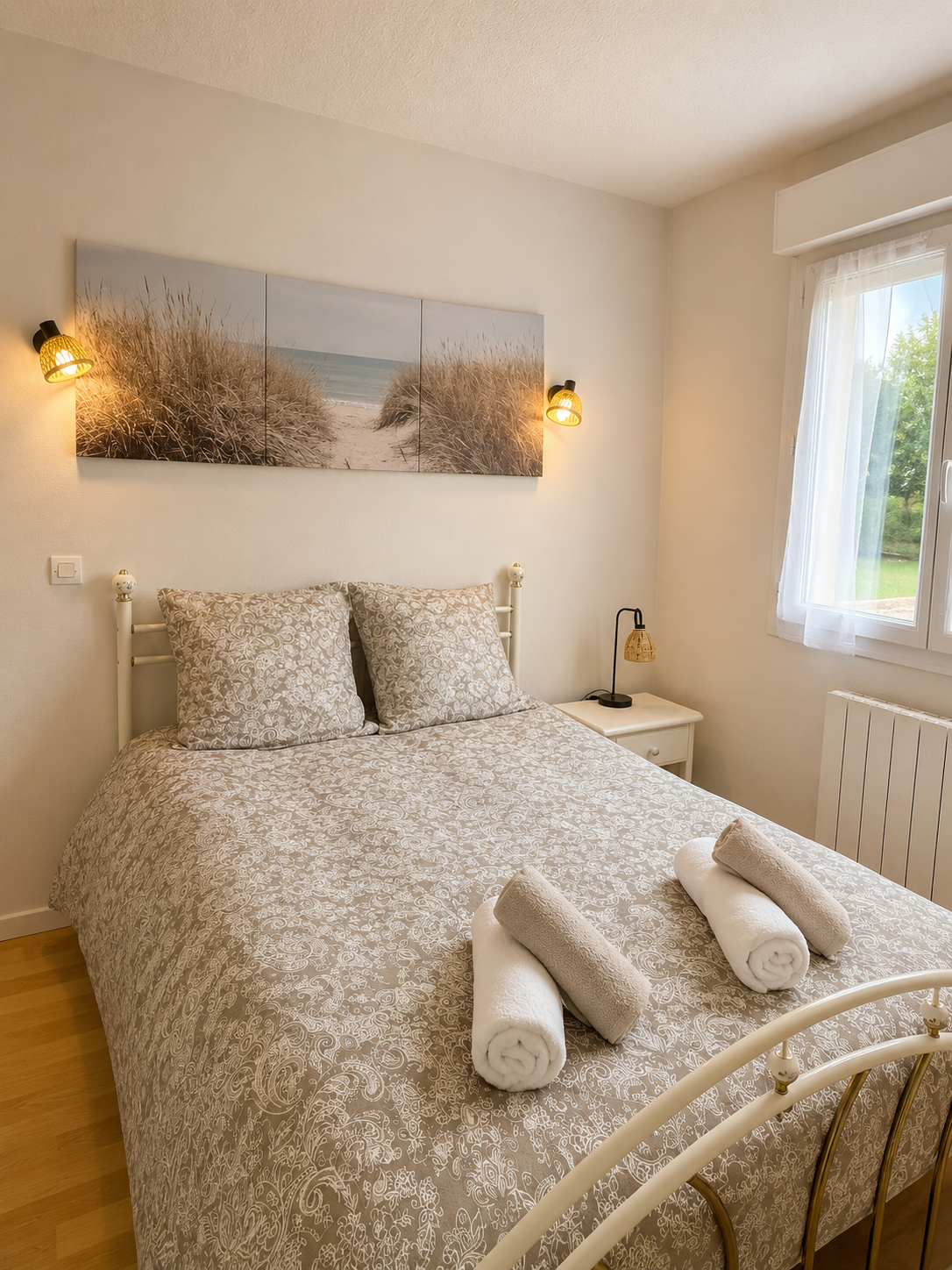Chambre avec lit double, draps beige à motifs, deux serviettes roulées sur le lit, tableau de plage avec dunes et mer, deux lampes de chaque côté du tableau, table de nuit avec lampe, fenêtre avec vue sur la nature