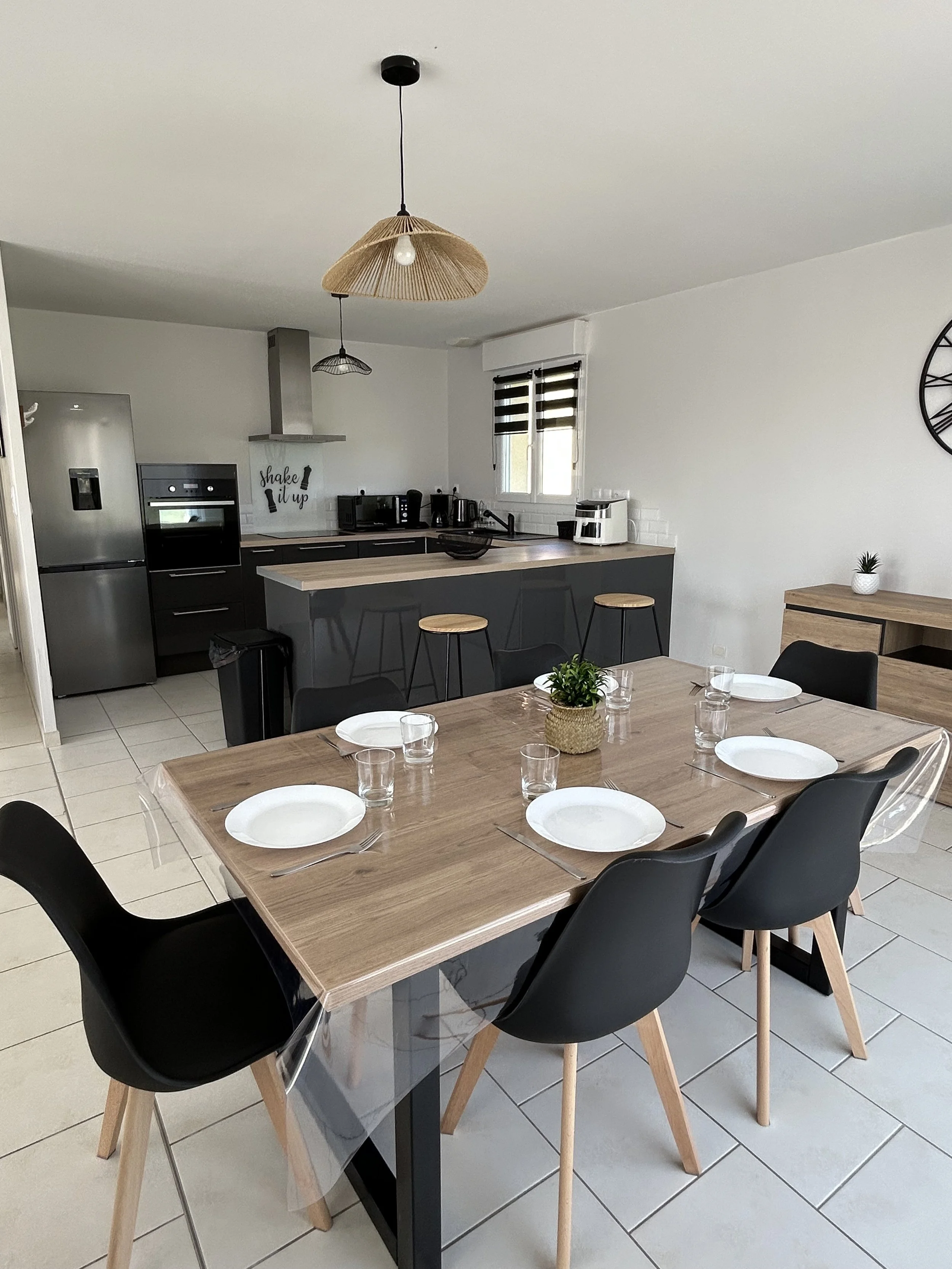 Salle à manger avec une table en bois, six chaises noires, assiettes et verres, et une cuisine en arrière-plan avec un plan de travail en bois, des appareils électroménagers, un mur blanc et deux fenêtres avec stores noirs