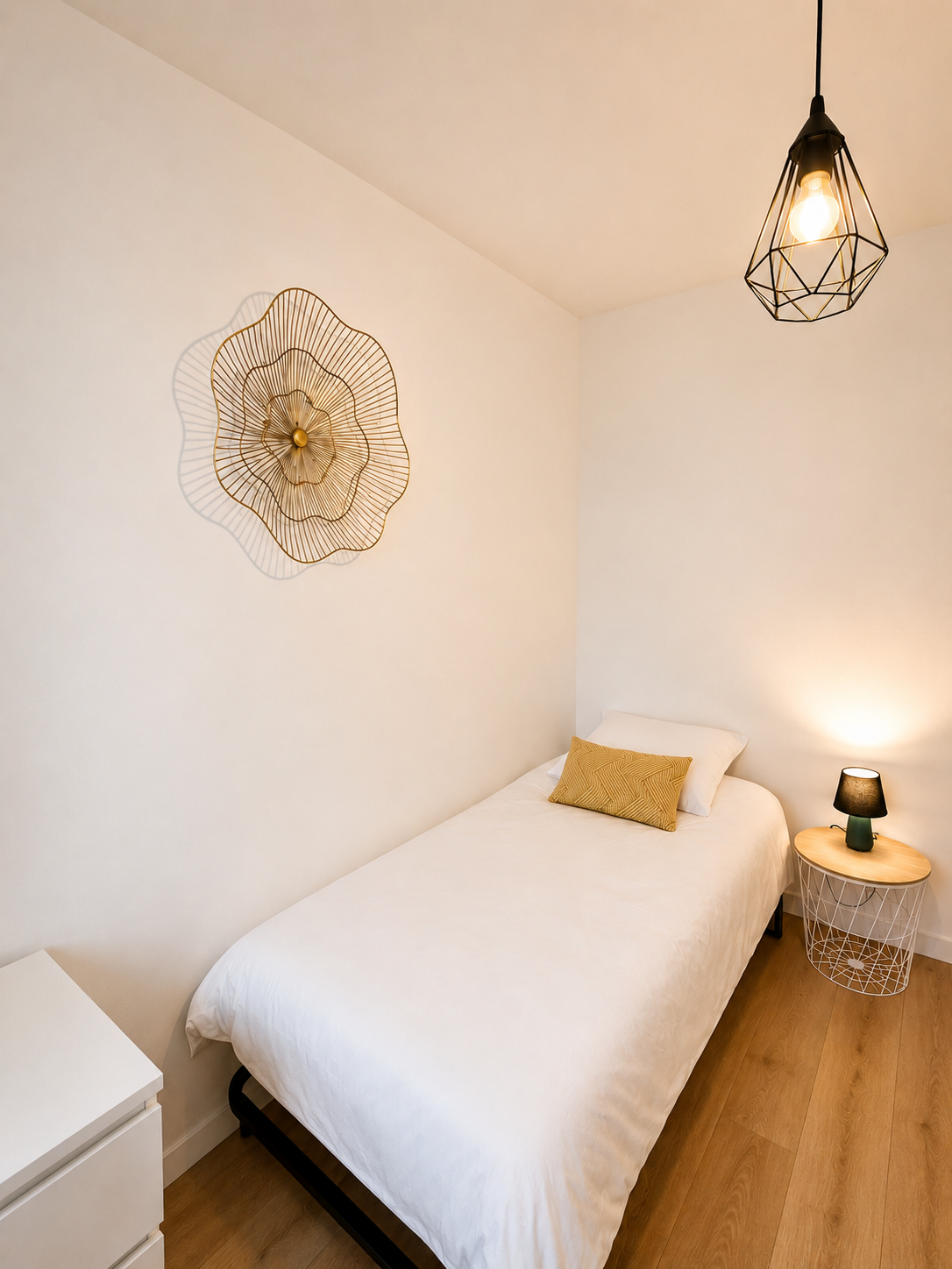 Chambre minimaliste avec un lit simple, une lampe de chevet, un tableau décoratif au mur et un luminaire suspendu.