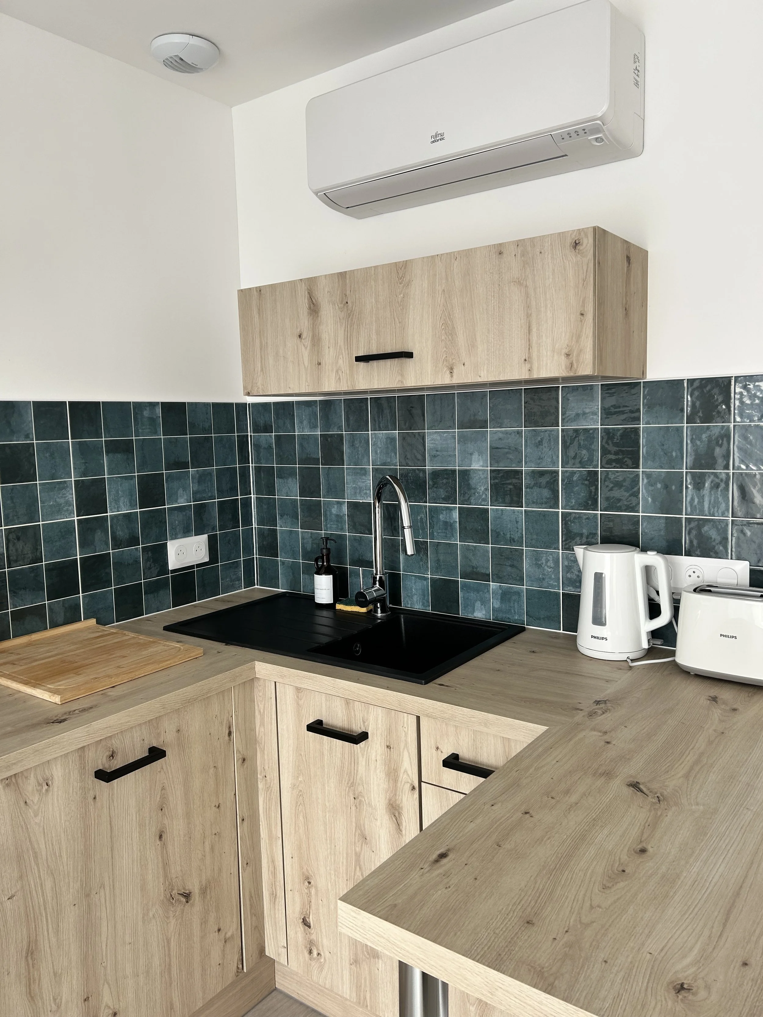 Coin de cuisine avec plan de travail en bois clair, évier avec robinet, derrière un mur de carrelage vert et quelques appareils électriques blancs.