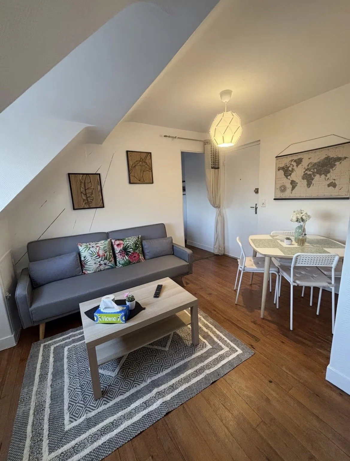 Un salon avec un canapé gris, une table basse, un tapis géométrique, une table à manger avec des chaises, un papier peint avec une carte du monde, des œuvres d'art sur le mur, et un lampadaire suspendu chaleureux.