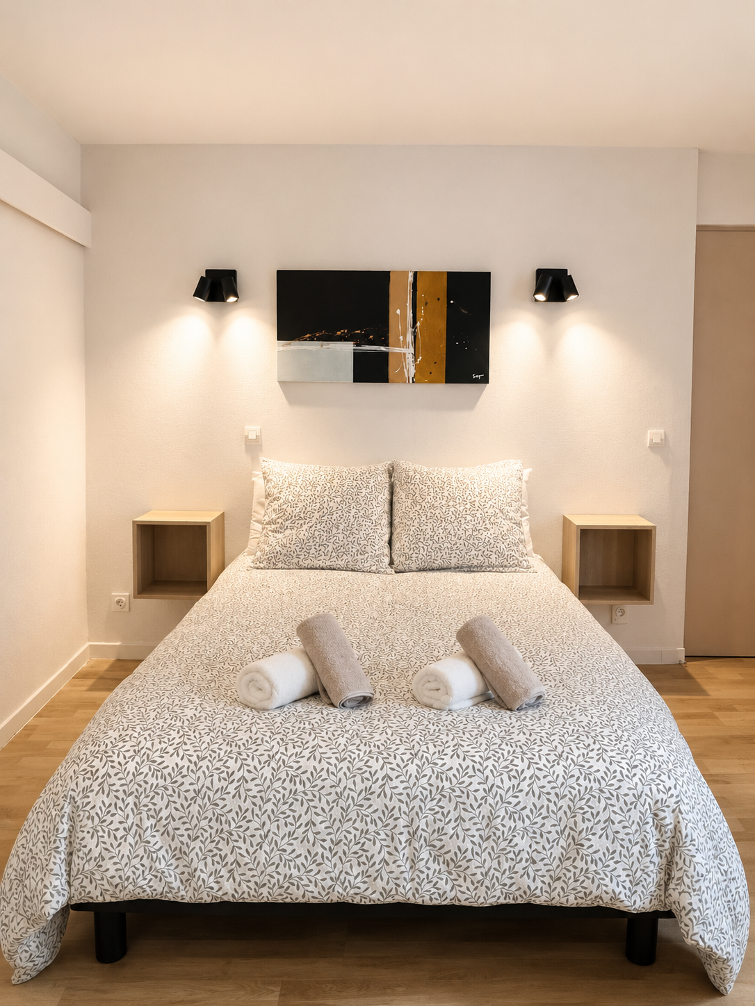 Chambre simple avec lit double, draps à motif de feuilles, deux oreillers, rouleaux de serviettes blancs et gris sur le lit, deux tables de nuit en bois clair, peinture abstraite noire, dorée et blanche au mur, deux appliques lumineuses noires.