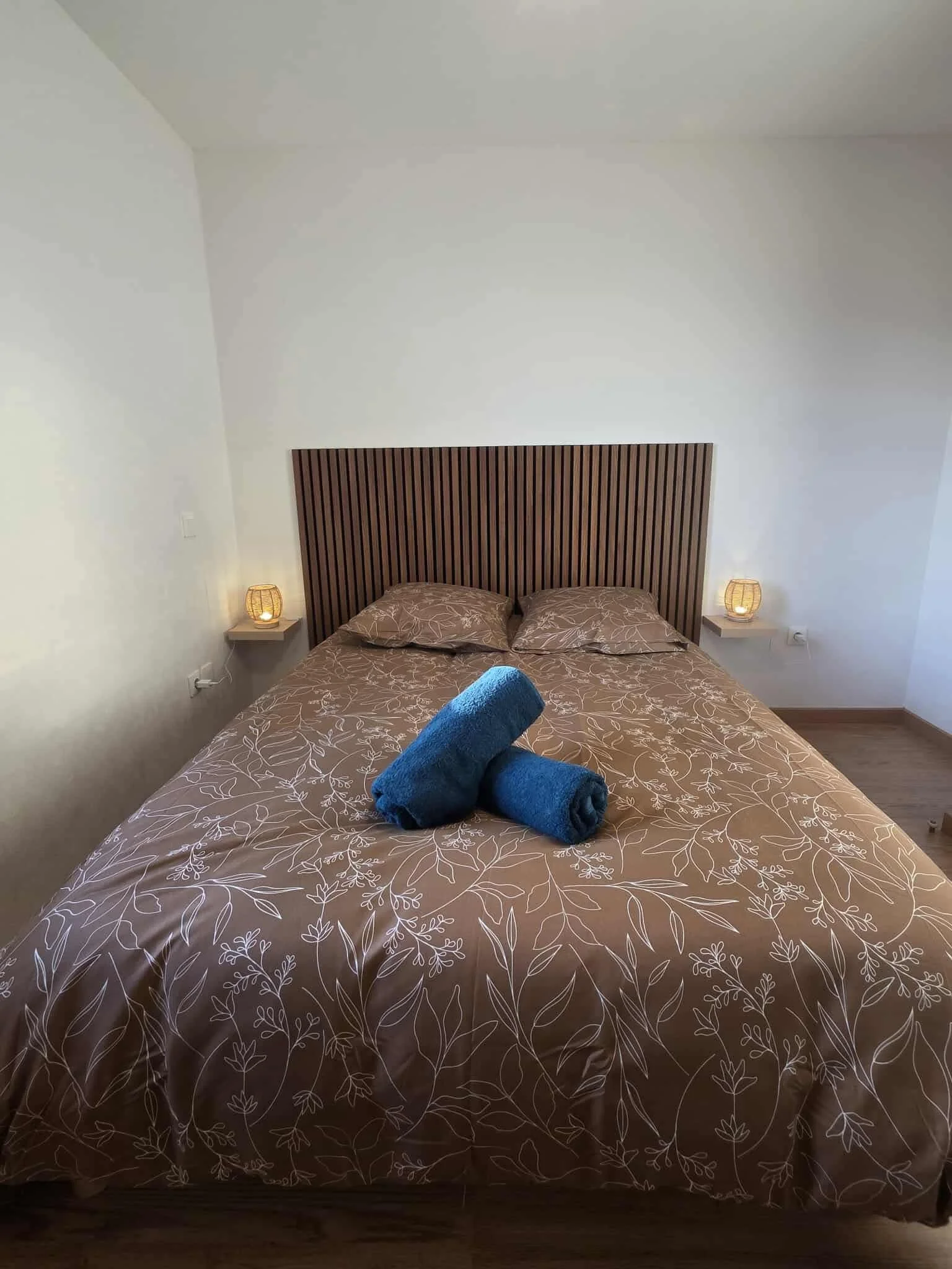 Un lit avec une couverture beige à motif floral, deux serviettes bleues roulées posées dessus, deux lampes de chevet avec lumières tamisées de chaque côté, un headboard en bois foncé en panneau vertical, murs blancs, sol en bois.