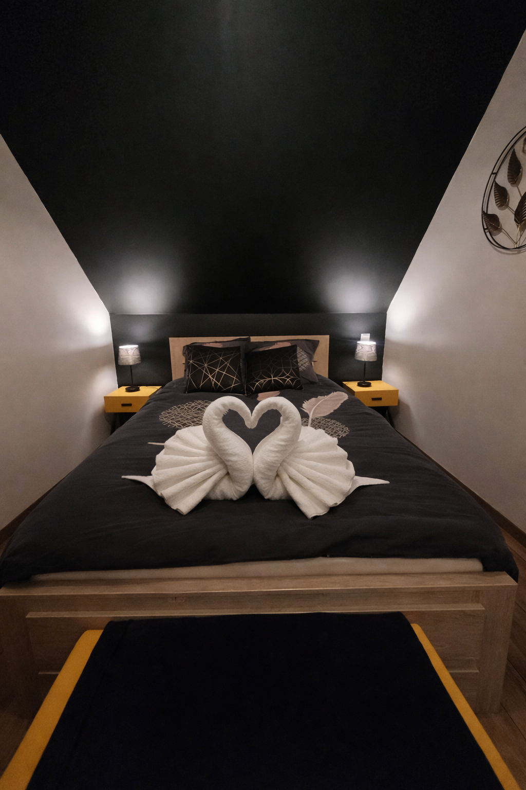 Chambre moderne avec un lit double, draps noirs et oreillers décoratifs, deux lampes de chevet, un lit plié en forme de cœur avec deux serviettes en forme de cygnes, et un mur noir au plafond.