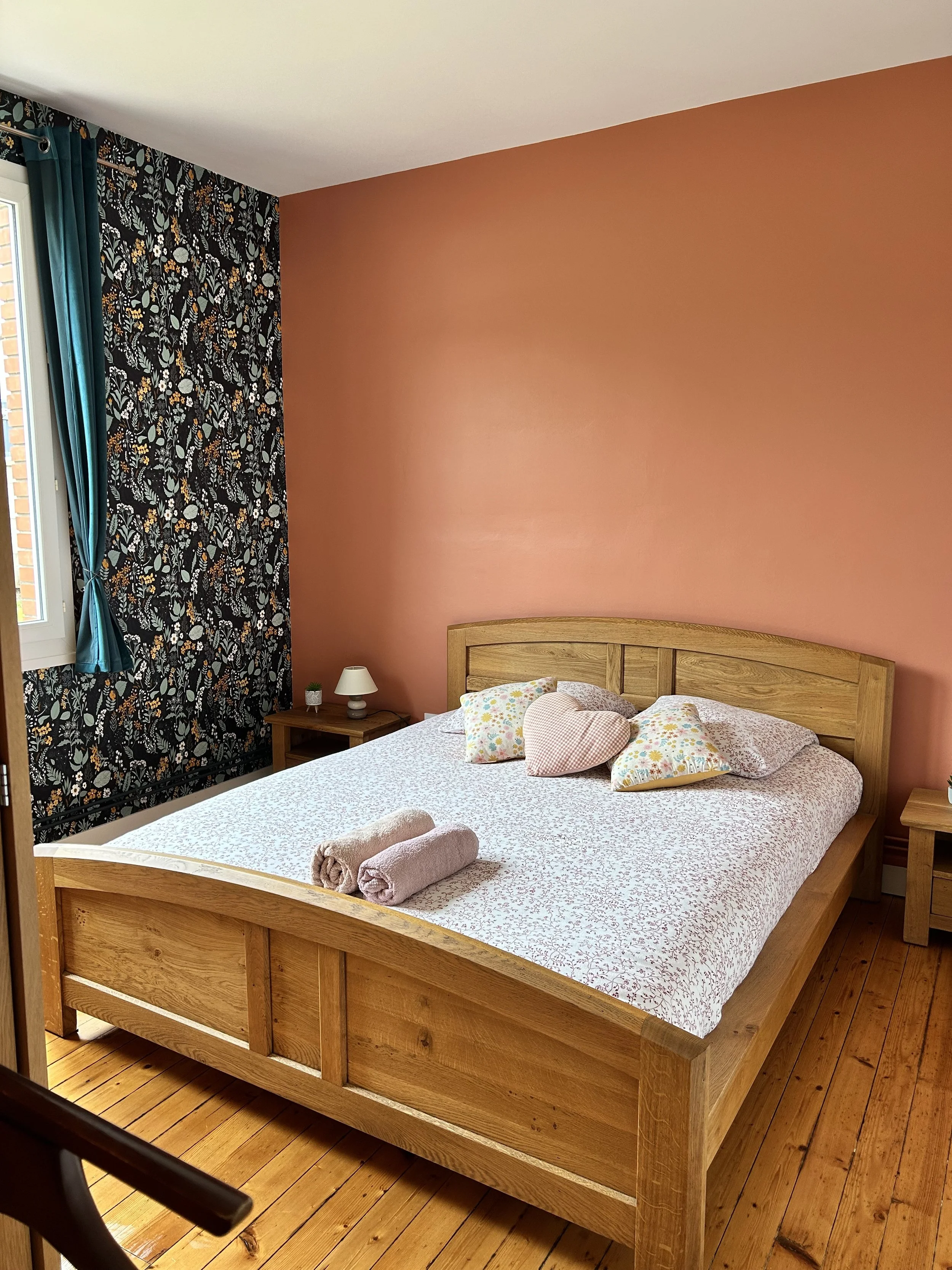 Chambre avec un lit en bois, des oreillers à motifs, deux serviettes roulées sur le lit, une table de chevet avec une petite lampe et une petite plante, une fenêtre avec des rideaux bleus et un mur peint en rose.