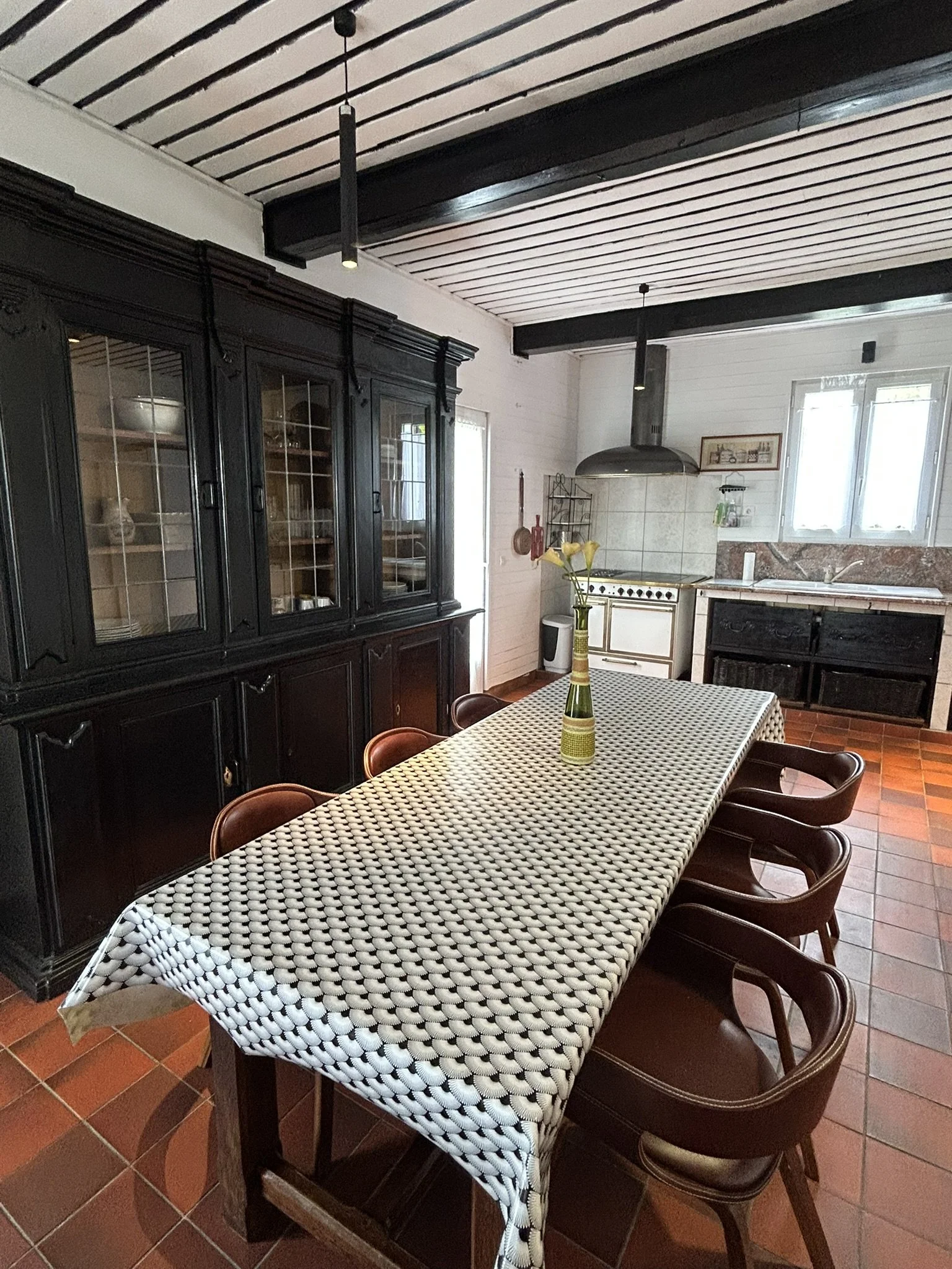Une salle à manger avec une table recouverte d'une nappe à motif tissé noir et blanc, entourée de plusieurs chaises en bois sombres. Sur la table se trouve un vase avec des fleurs jaunes. À l'arrière, une cuisine avec des meubles sombres, un évier sous une fenêtre, et un plan de travail en granite. Le plafond est en bois avec des poutres apparentes, et il y a deux luminaires suspendus.