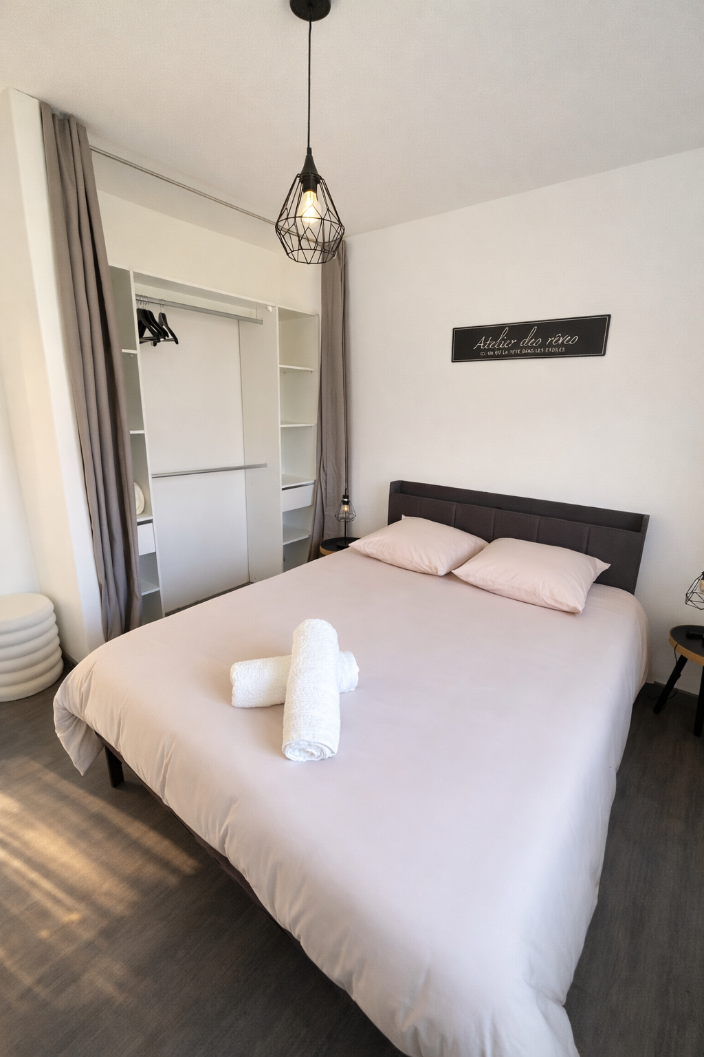 Chambre moderne avec lit double, draps roses, deux serviettes blanches roulées sur le lit, lampes de chevet, murs blancs, porte-miroir avec placard intégré, décor minimaliste et suspensions en métal