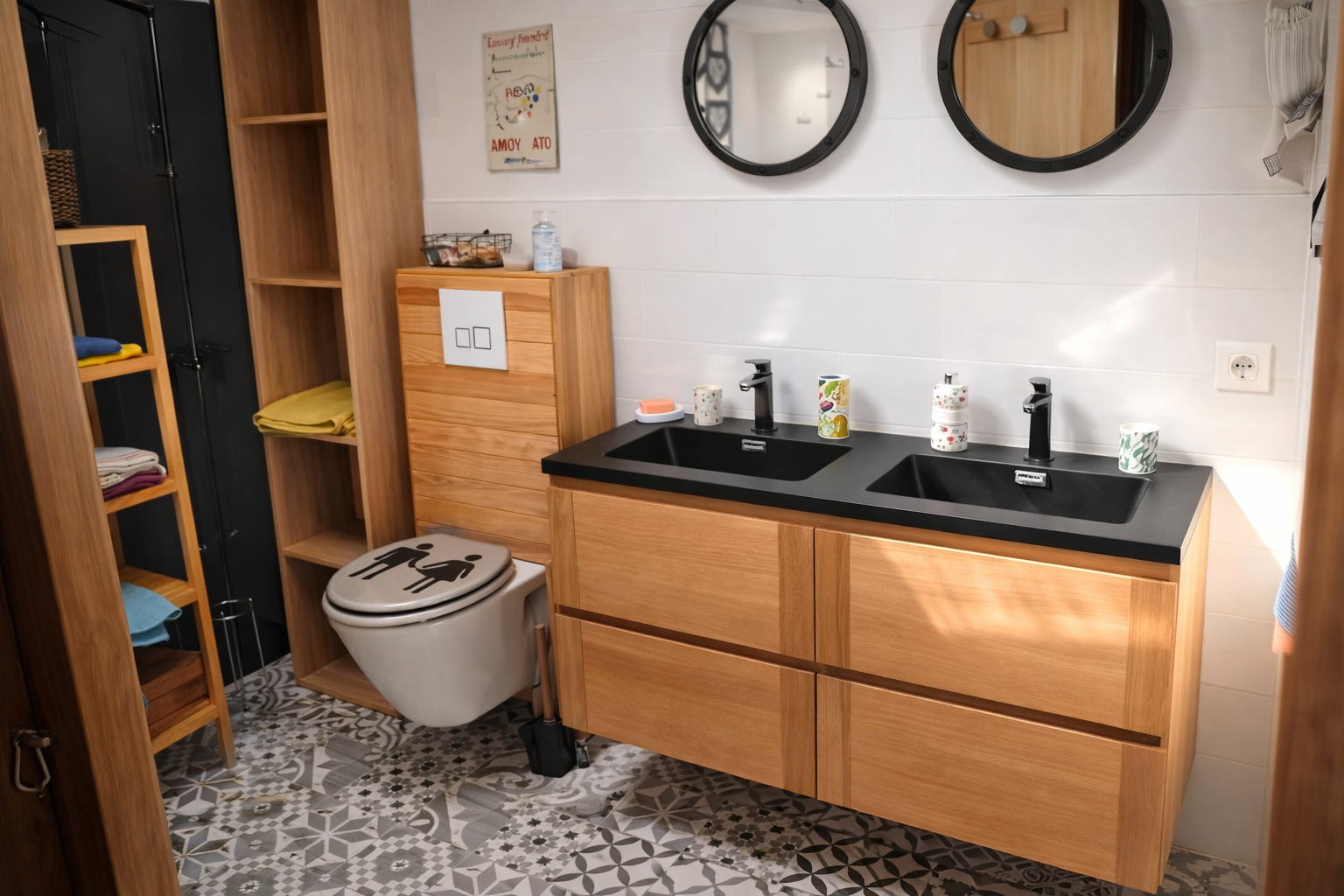 Salle de bain avec lavabo double, miroir rond, toilettes, étagères en bois avec serviettes, décor coloré, plancher en carrelage géométrique.
