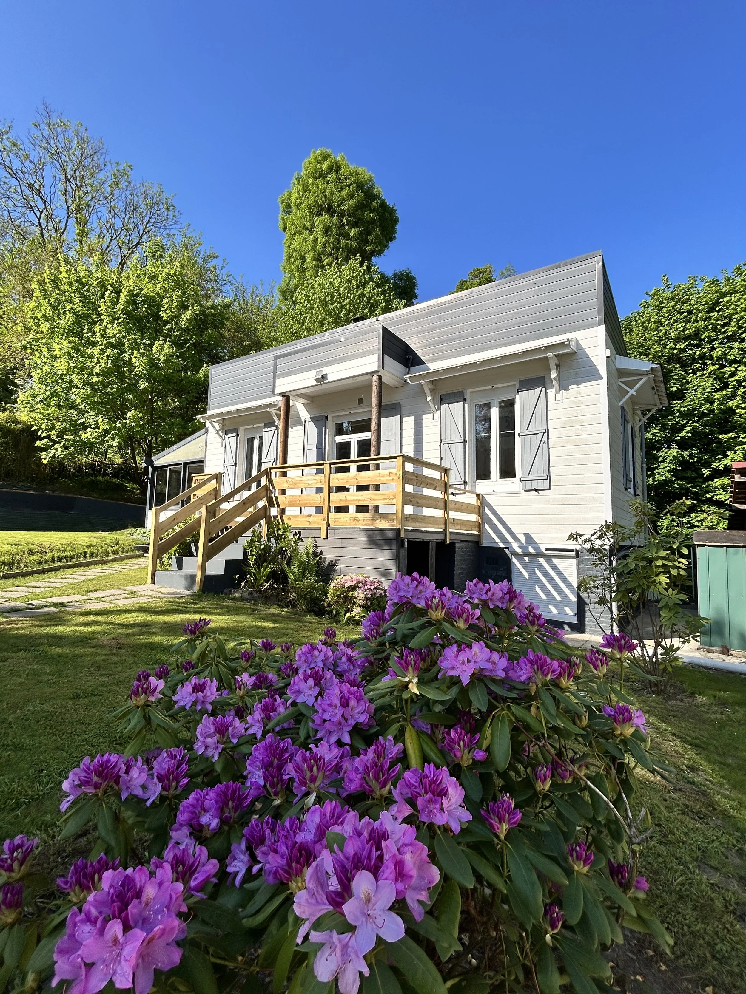 Jardin avec des fleurs violettes devant une maison en bois blanc avec gonds à volets gris et rampes en bois, entourée d'arbres verts, sous un ciel bleu clair.
