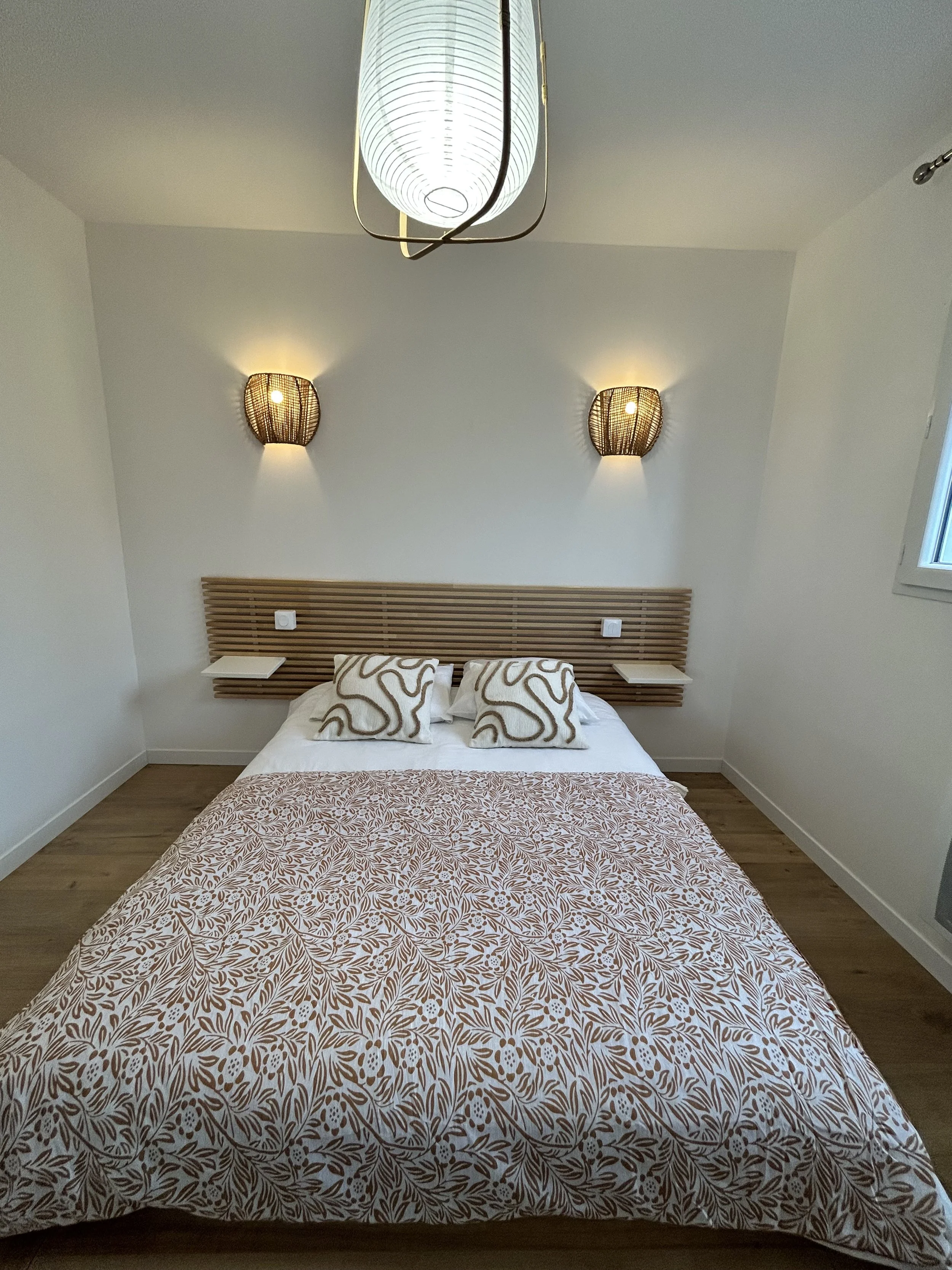 Lit simple dans une chambre moderne avec tête de lit en bois, deux lampes murales en rotin de chaque côté et deux coussins avec un motif abstrait sur le lit, sous un plafonnier blanc